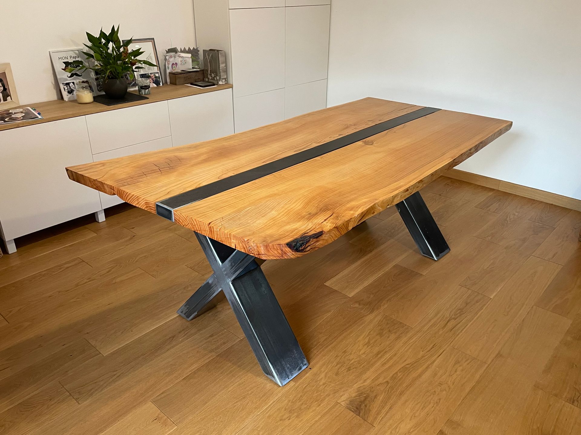 Une table de salle à manger en bois brut avec une bande centrale noire et des pieds en métal noir en forme de X, posée sur un sol en bois.