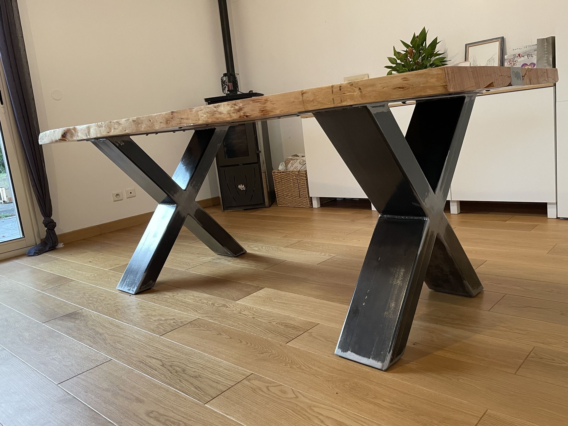 Une table à manger rustique avec un plateau épais en bois brut et des pieds en métal noir en forme de X, dans un intérieur domestique.