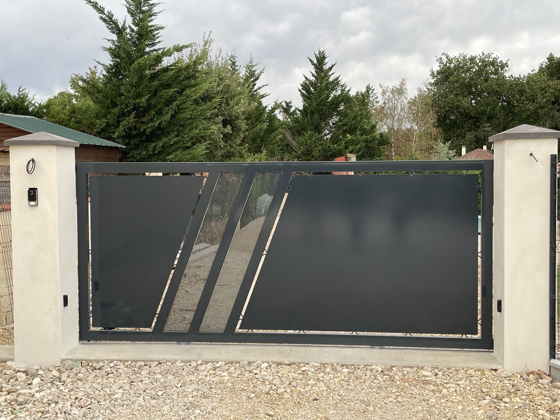 Portail coulissant moderne en métal gris foncé avec un motif géométrique ajouré décoratif, situé entre deux piliers beiges.