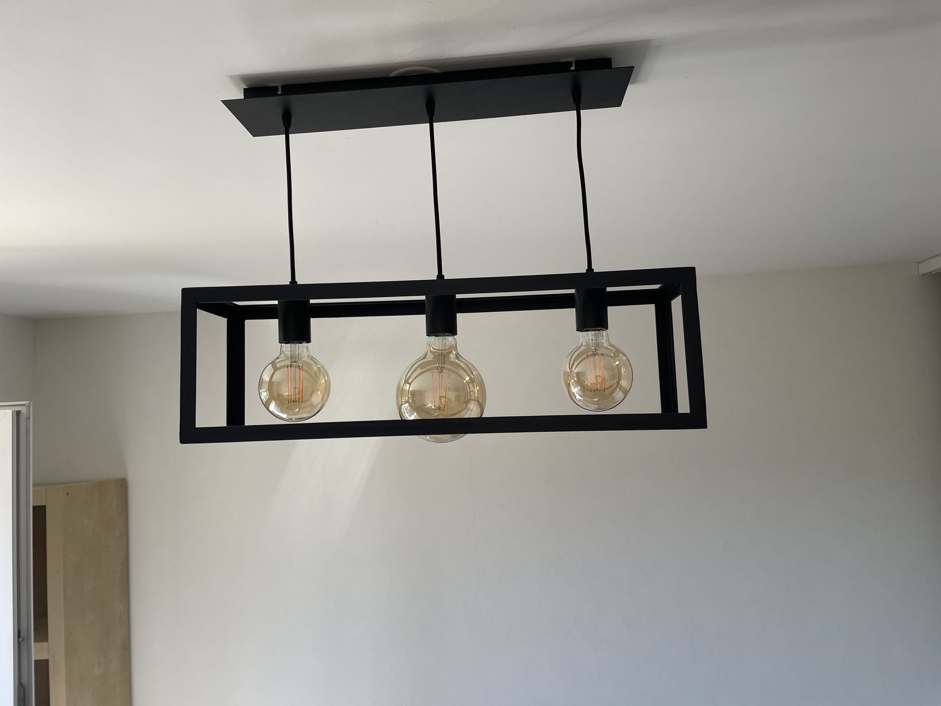 Un luminaire suspendu rectangulaire moderne en métal noir avec trois ampoules apparentes de style Edison, suspendu au plafond.