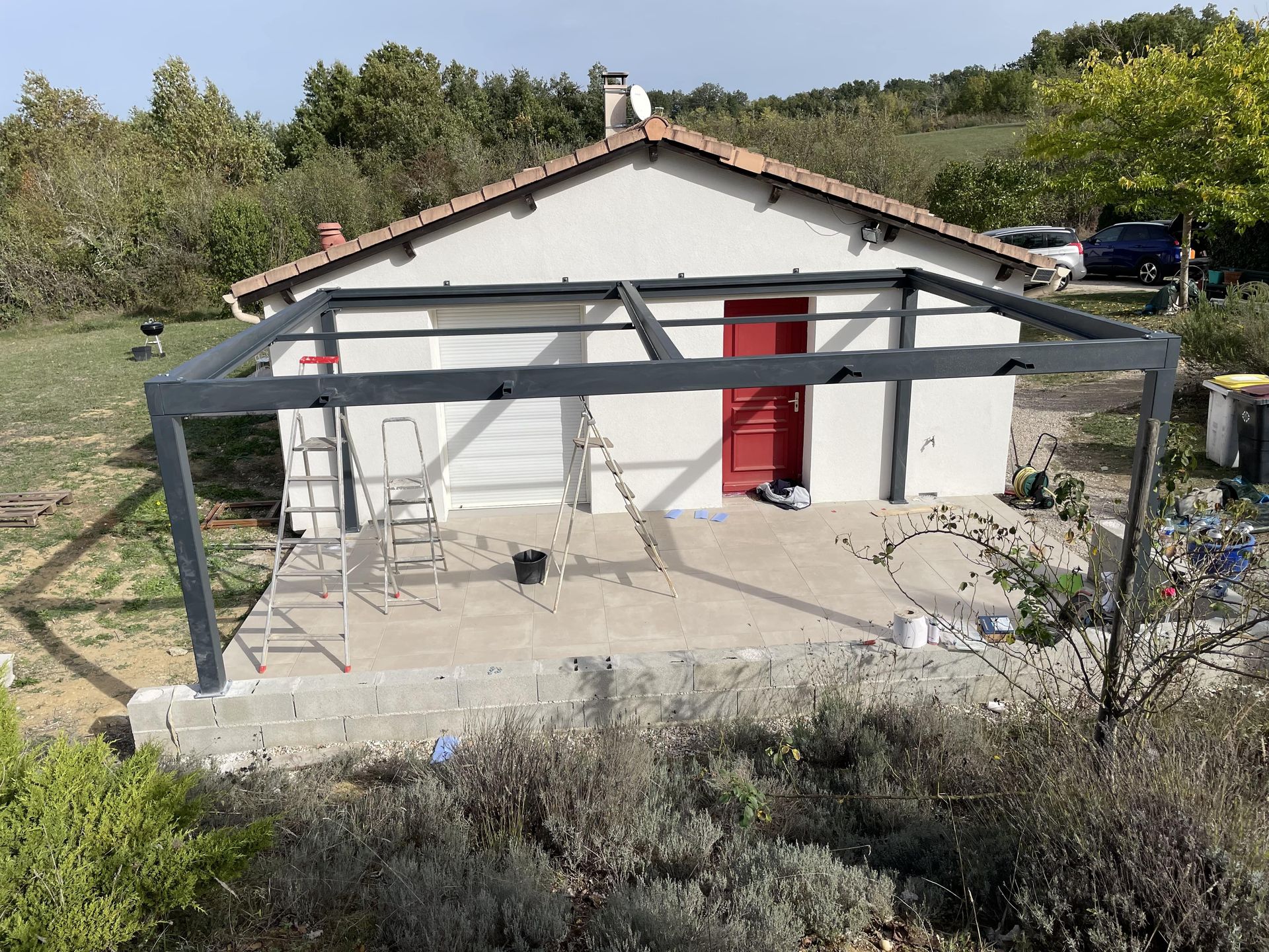 Une pergola en métal gris foncé se dresse sur une terrasse en pierre devant une maison blanche à la porte rouge et au toit de tuiles.