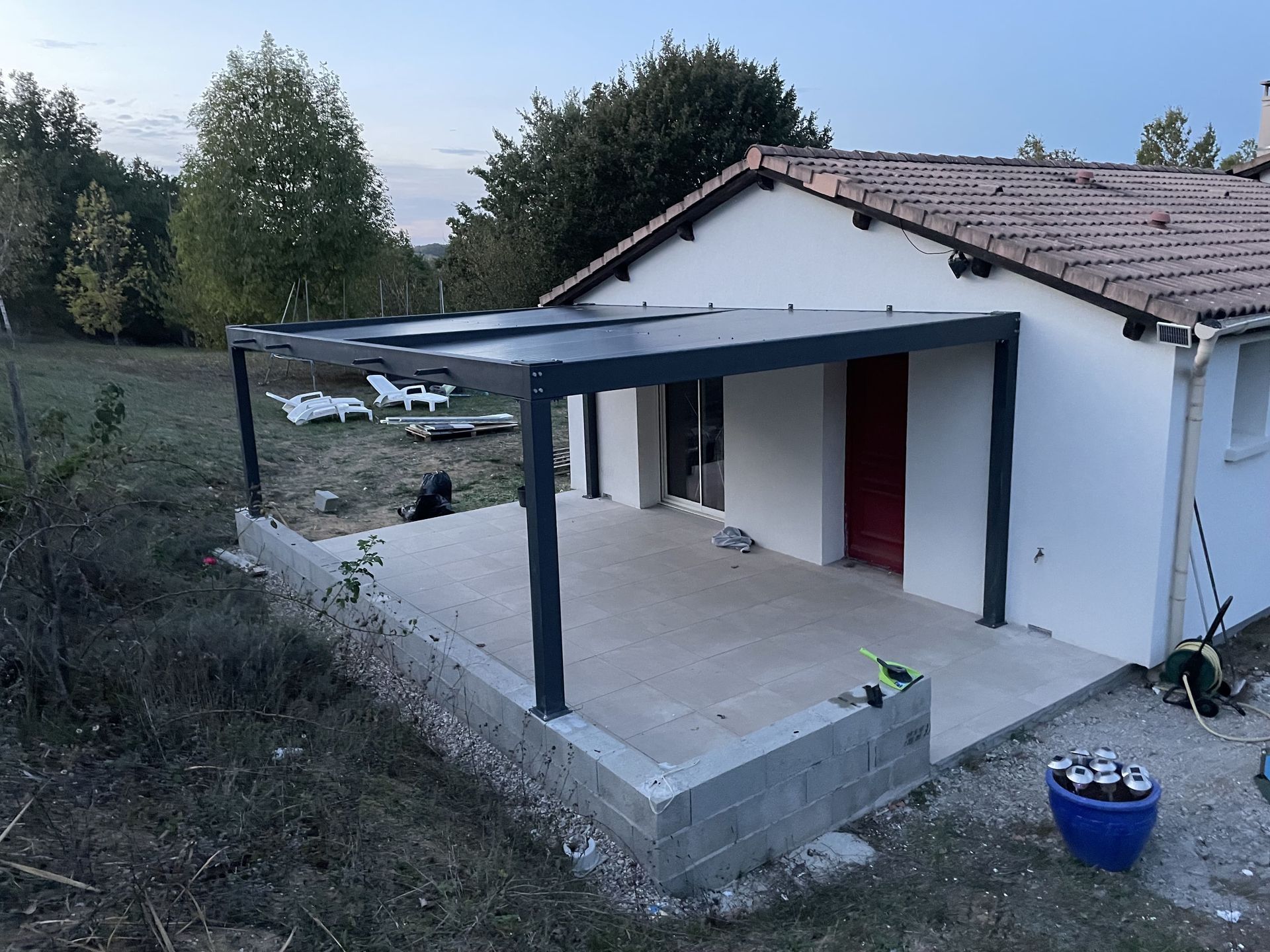 Une pergola moderne gris foncé installée au-dessus d'une terrasse en béton, à côté d'une maison blanche avec une porte rouge.