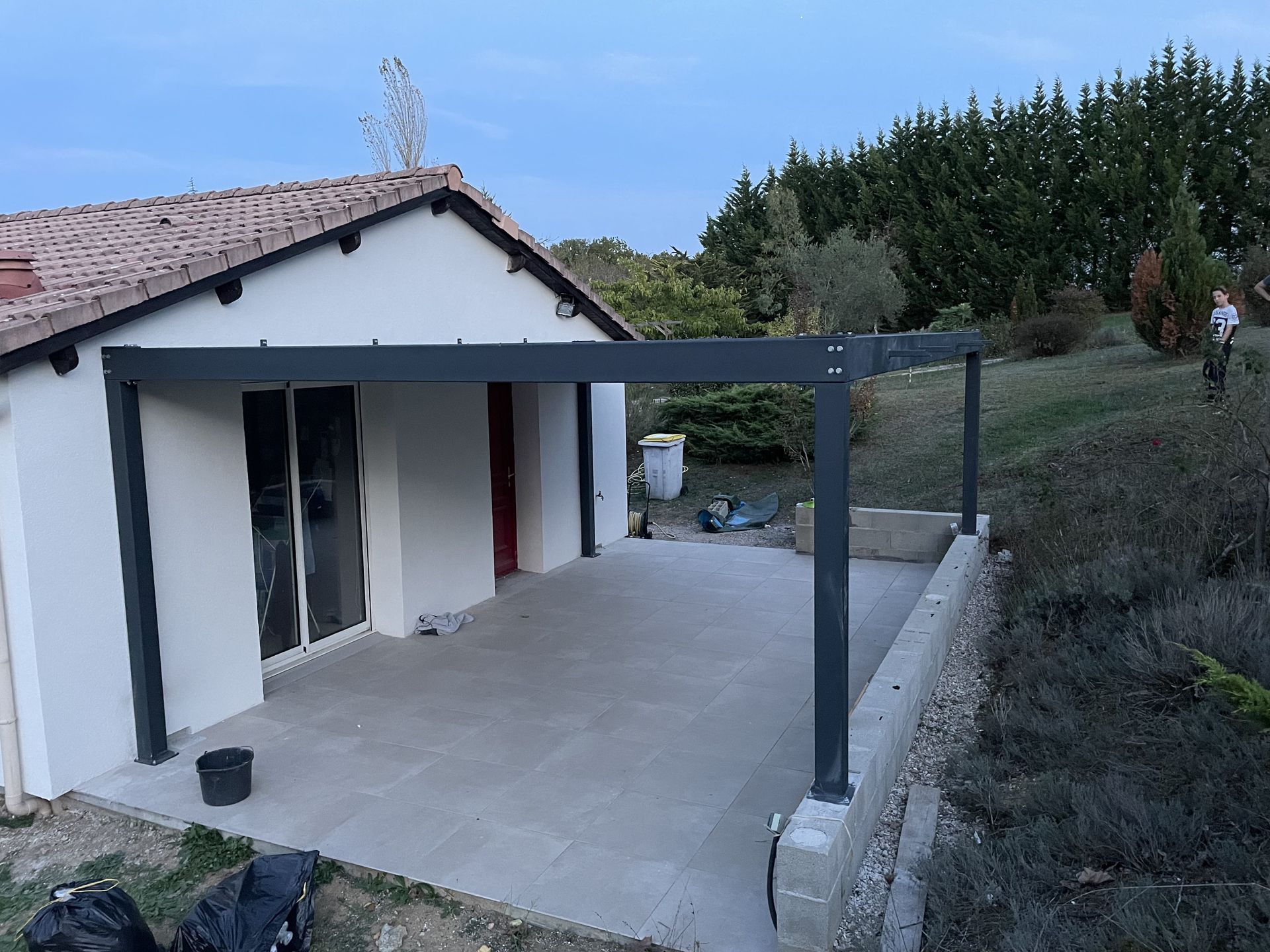 Une maison avec une pergola en métal gris foncé récemment installée au-dessus d'une terrasse pavée de couleur claire.