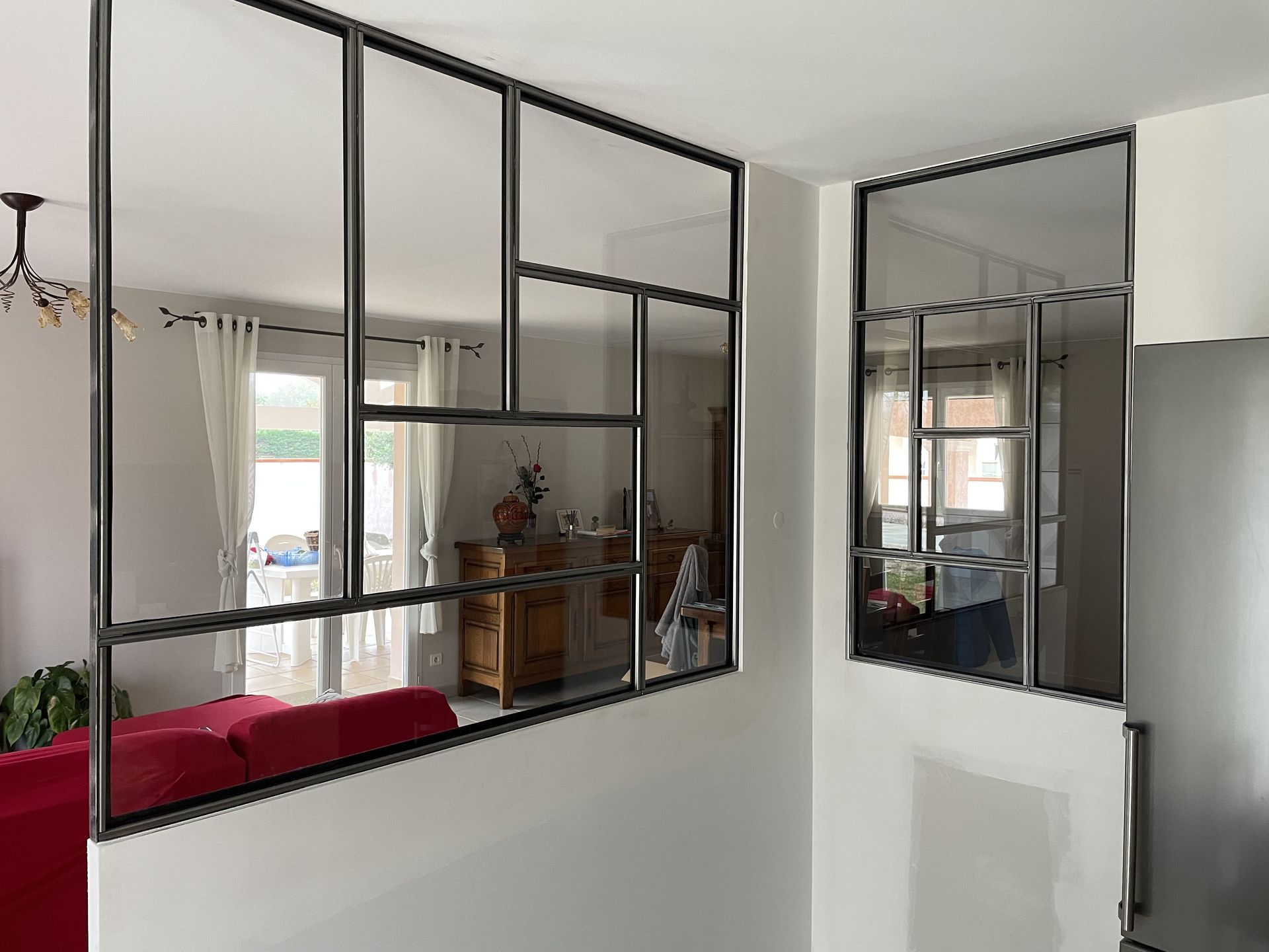 Des cloisons en verre à cadre métallique noir, fixées sur des demi-murs blancs, séparent un salon d'un espace adjacent.