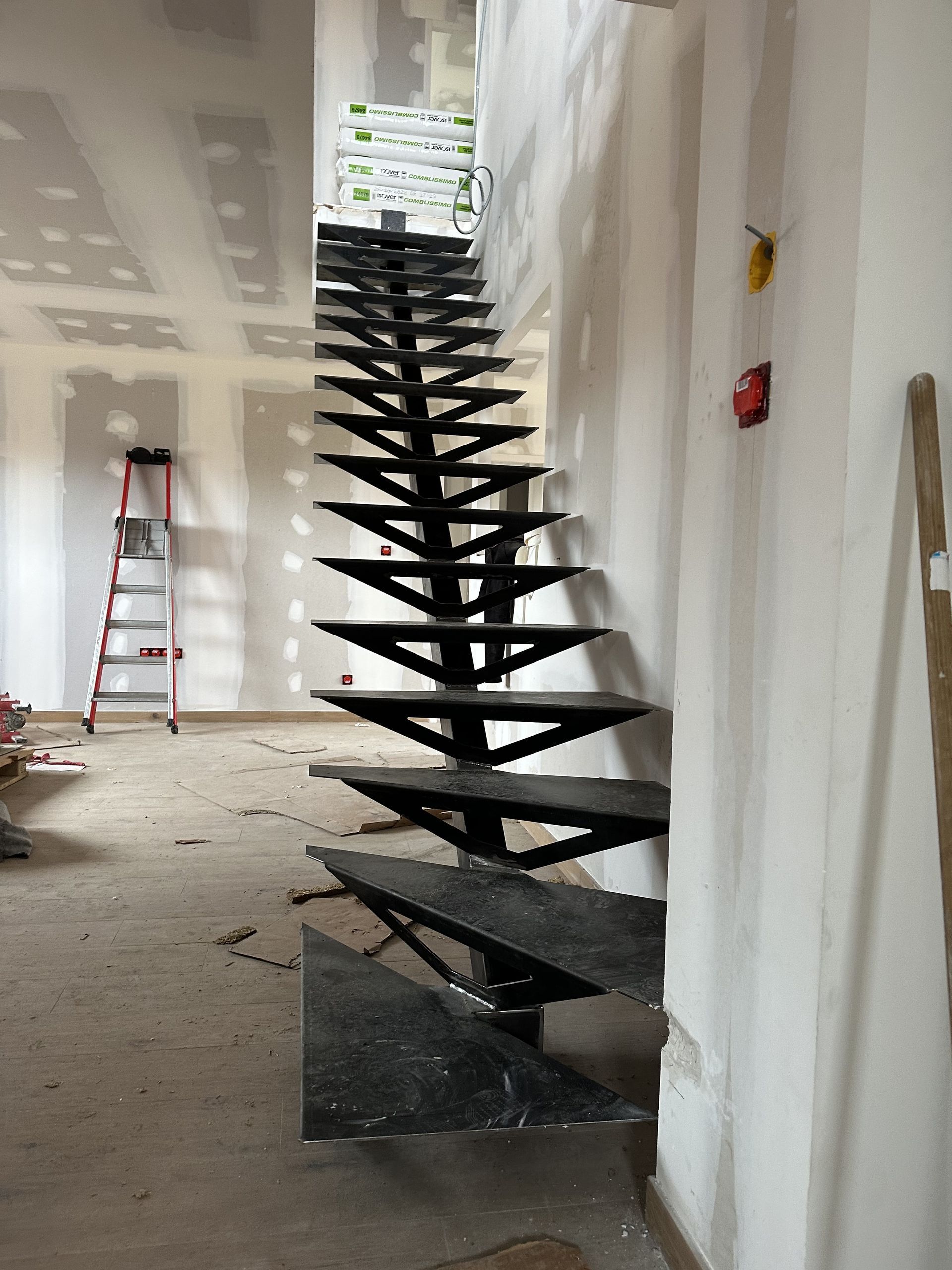 Un escalier moderne à limons ouverts, avec des marches triangulaires foncées, installé dans une pièce inachevée avec des cloisons sèches et une échelle.