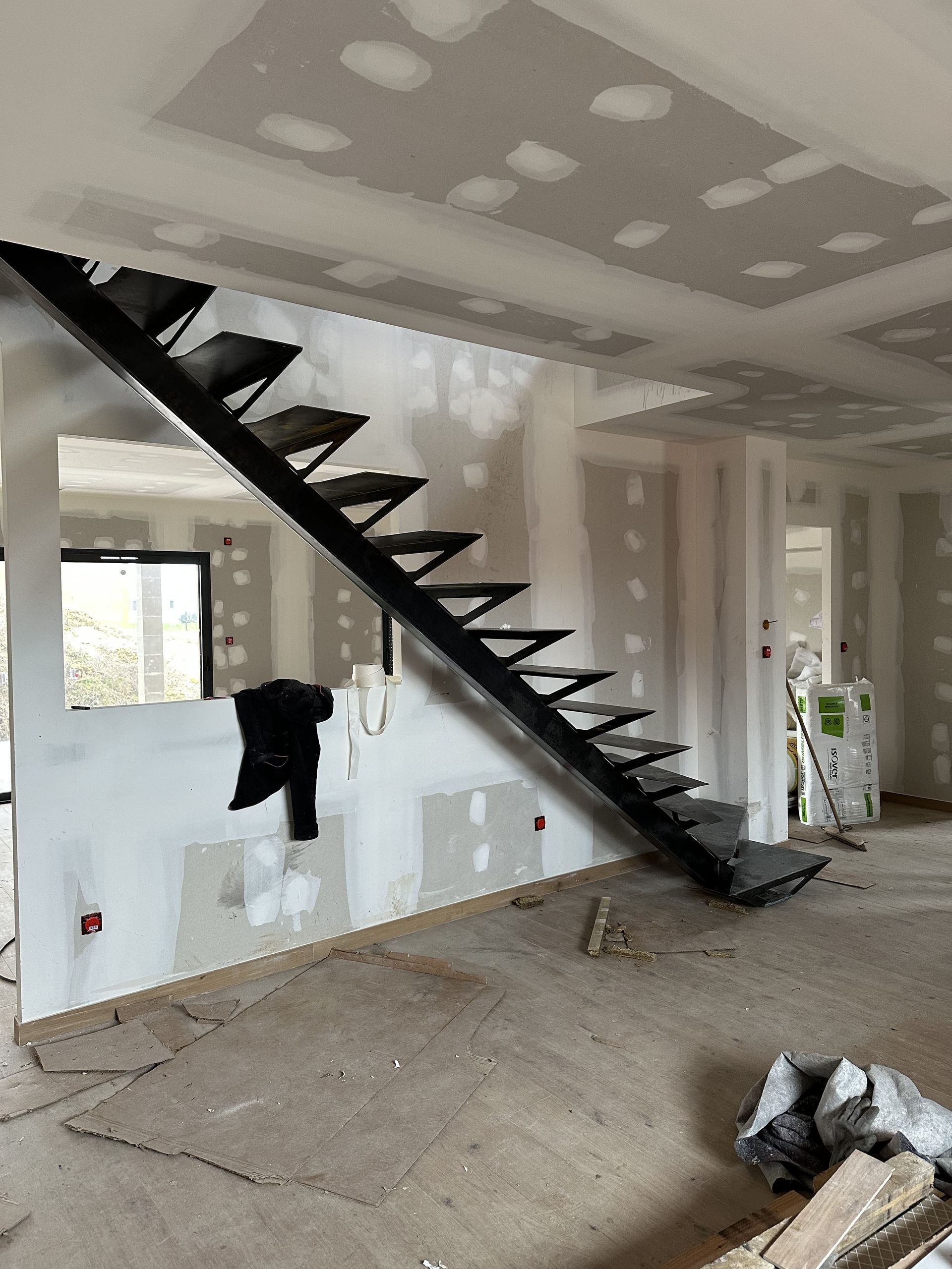 Une pièce inachevée présente une structure d'escalier en métal noir foncé fixée contre un mur aux panneaux de plaques de plâtre apparents.
