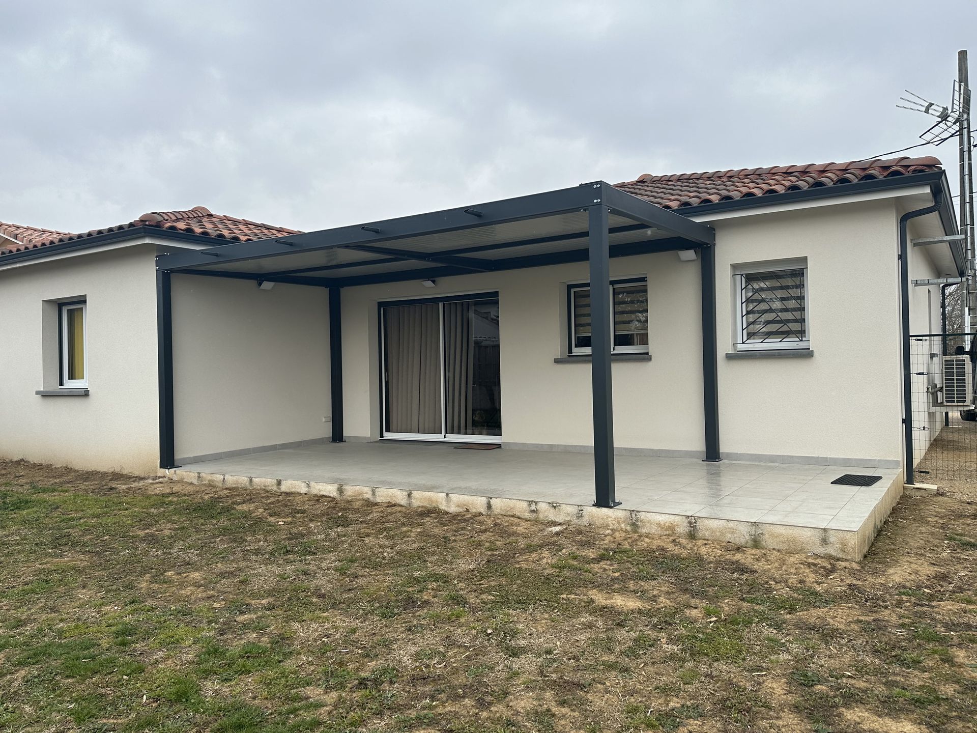 Une maison de plain-pied avec une façade beige, un toit de tuiles et une grande pergola en métal noir au-dessus d'une terrasse carrelée.