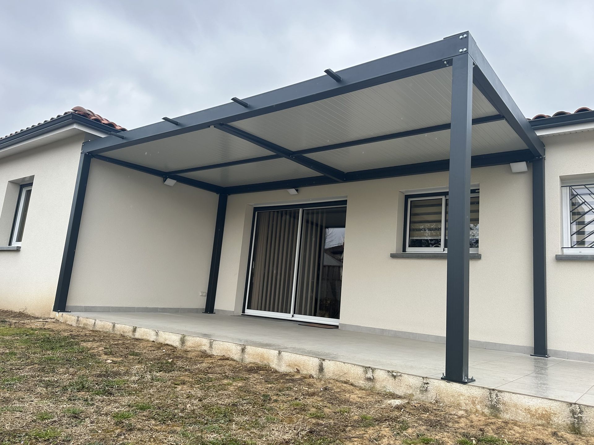 Une pergola moderne en métal gris foncé, fixée à l'arrière d'une maison beige avec une terrasse en béton.