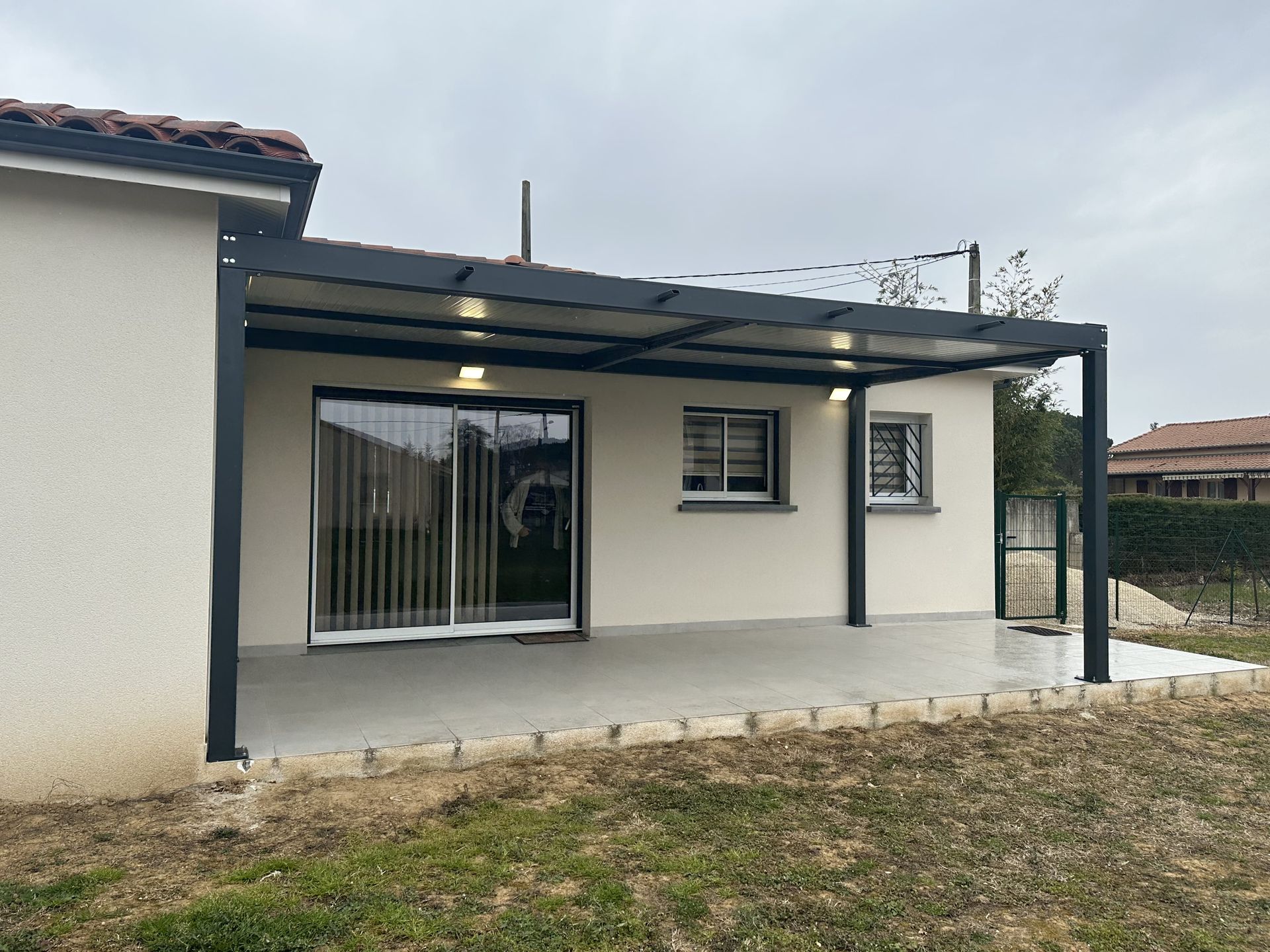 Une pergola moderne en métal noir, adossée à une maison beige, ombrage une terrasse en béton grâce à deux plafonniers lumineux.