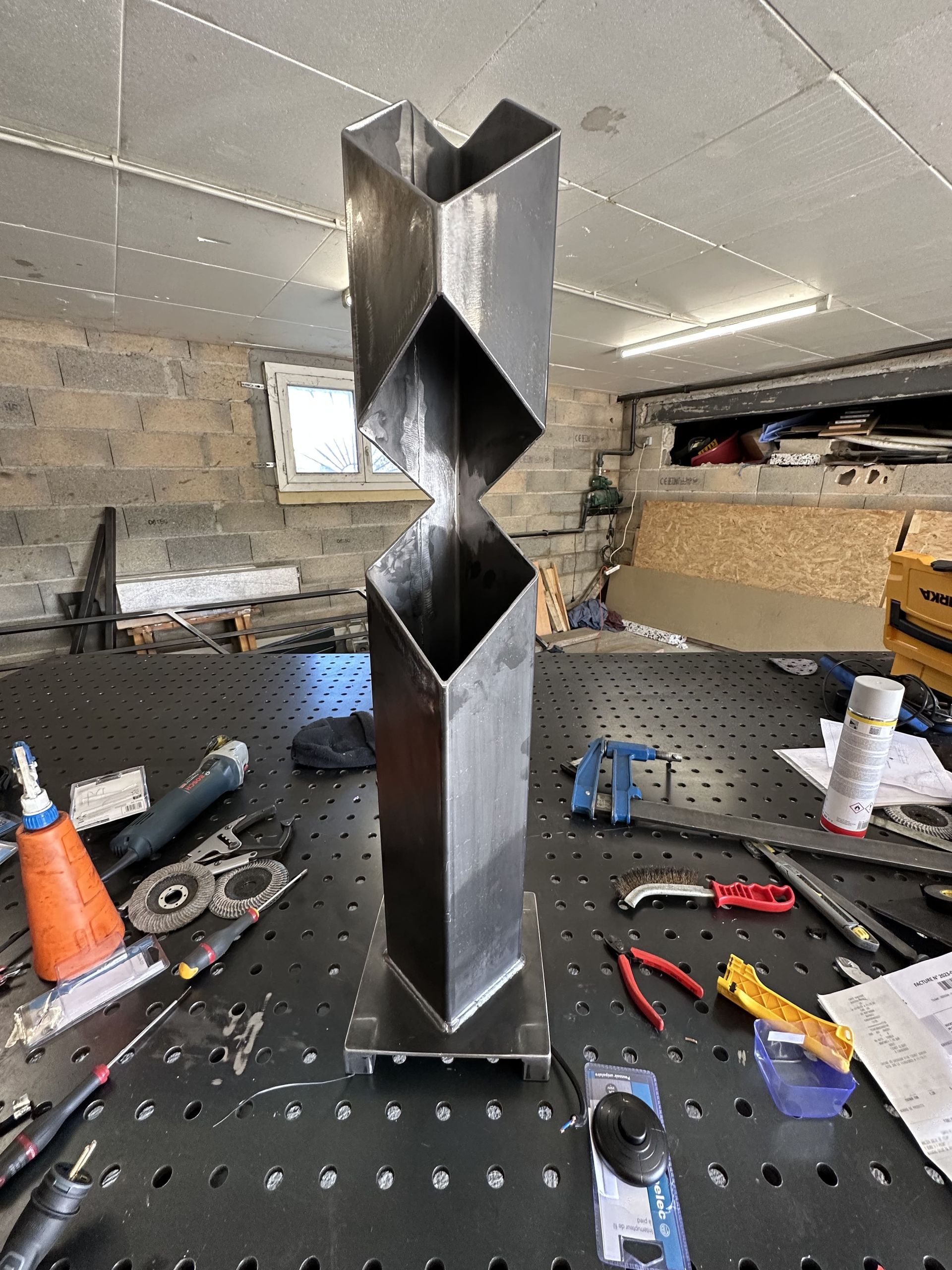 Une sculpture métallique verticale, composée de tubes carrés découpés en diagonale, posée sur un établi dans un atelier.