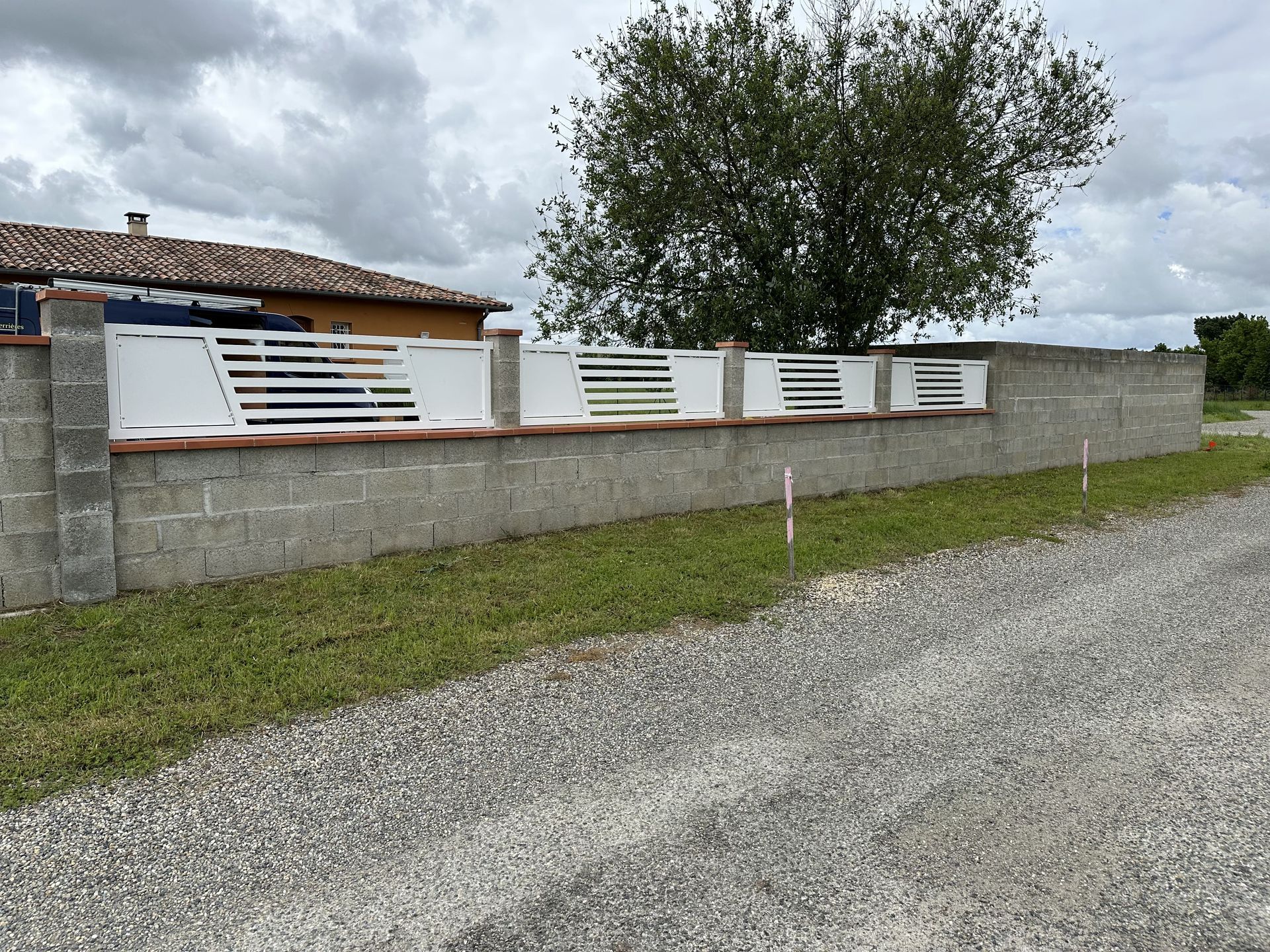 Un long mur de béton gris surmonté d'une clôture métallique blanche à lattes horizontales, situé le long d'un chemin de gravier près d'une maison.