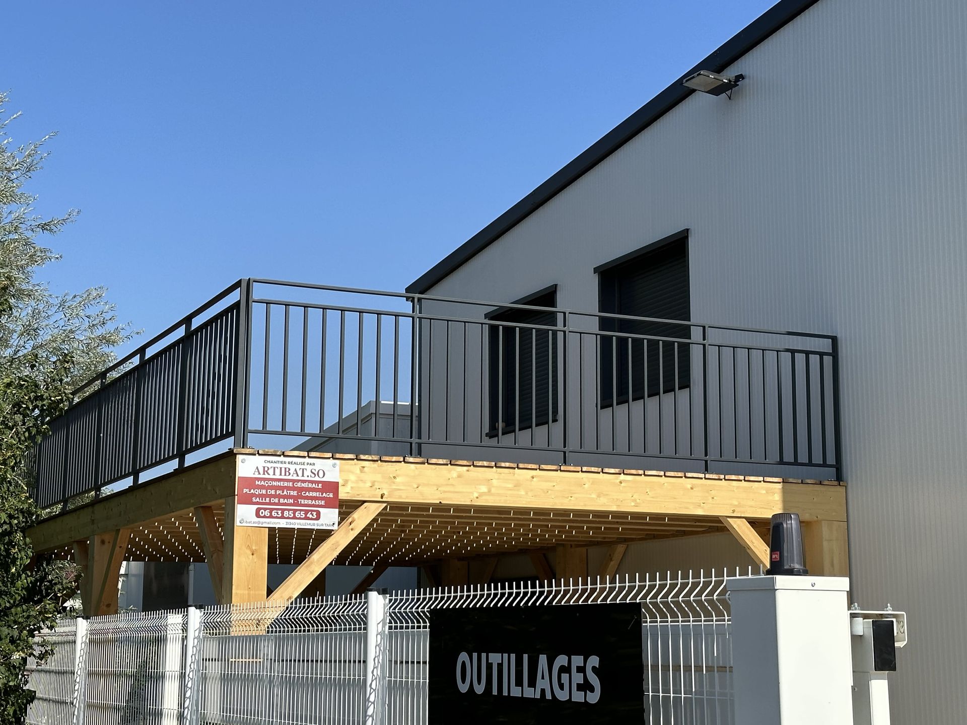 Une terrasse en bois avec une rambarde en métal noir fixée à un bâtiment, avec un panneau indiquant « OUTILLAGES » sur la clôture en contrebas.
