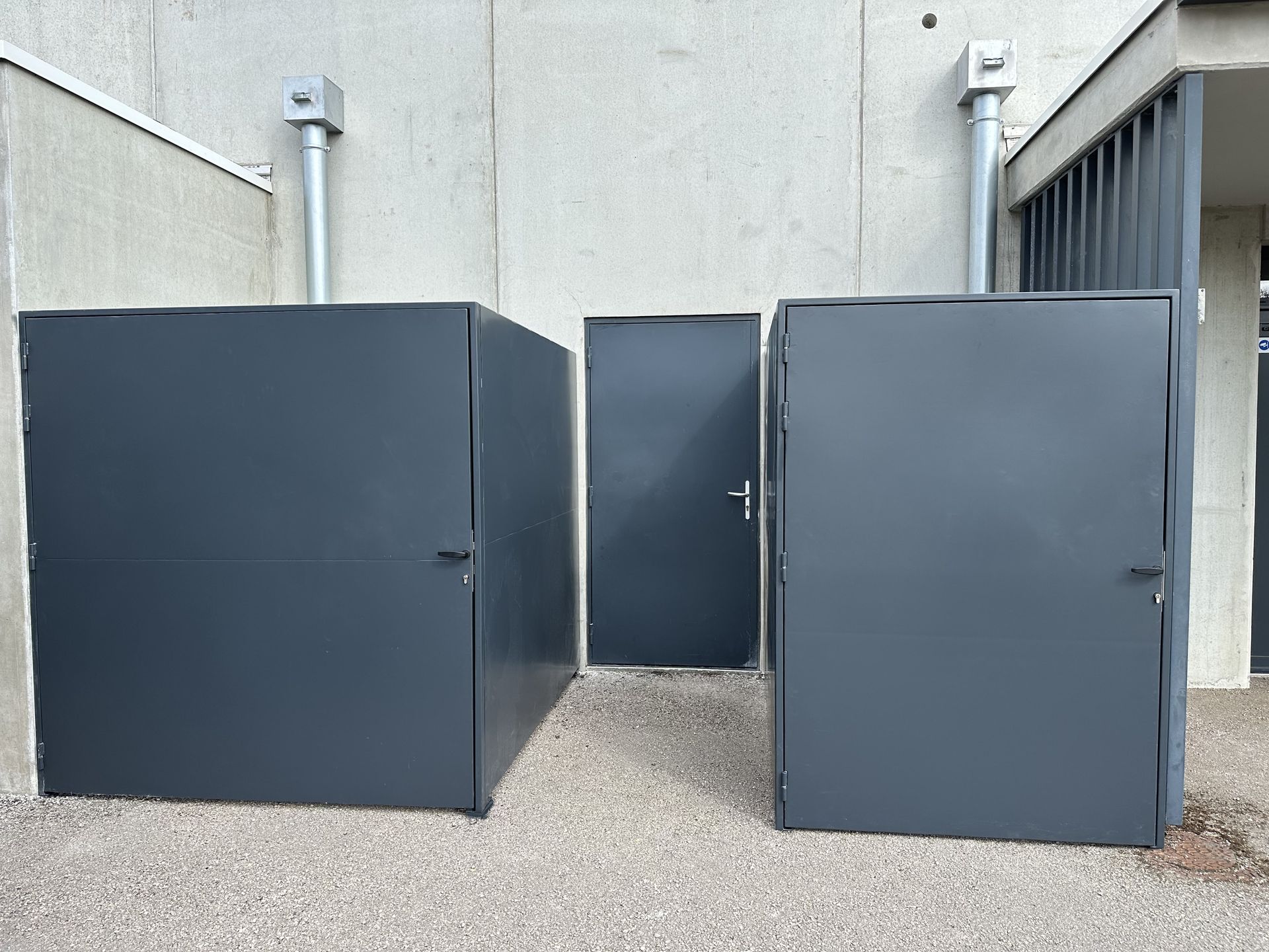 Deux unités de rangement en métal gris foncé et une porte d'accès assortie sont adossées à un mur en béton sur un sol gravillonné.