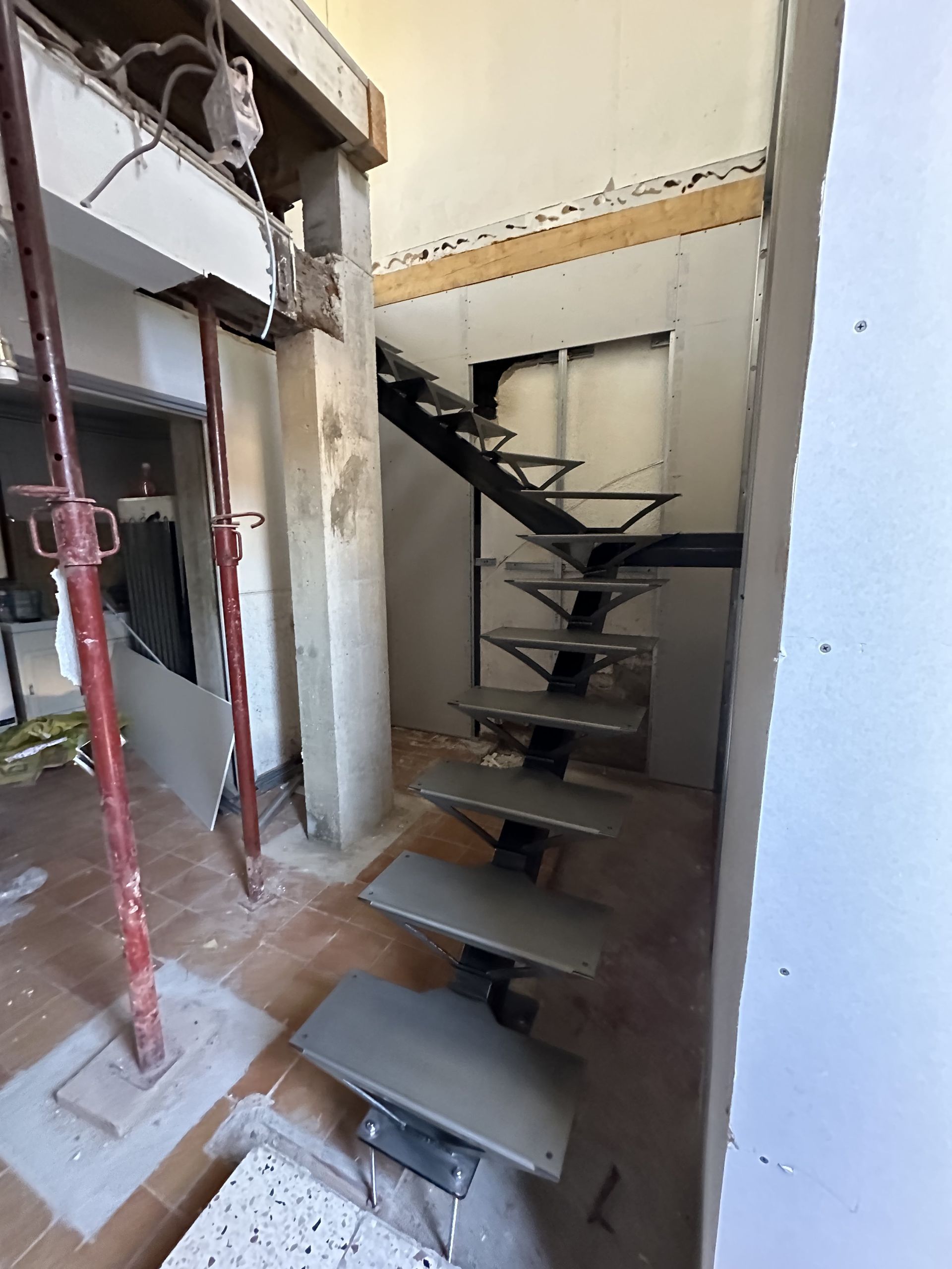 Un escalier aux marches métalliques grises en construction dans une pièce aux ossatures apparentes, aux vérins de soutien et aux murs non finis.