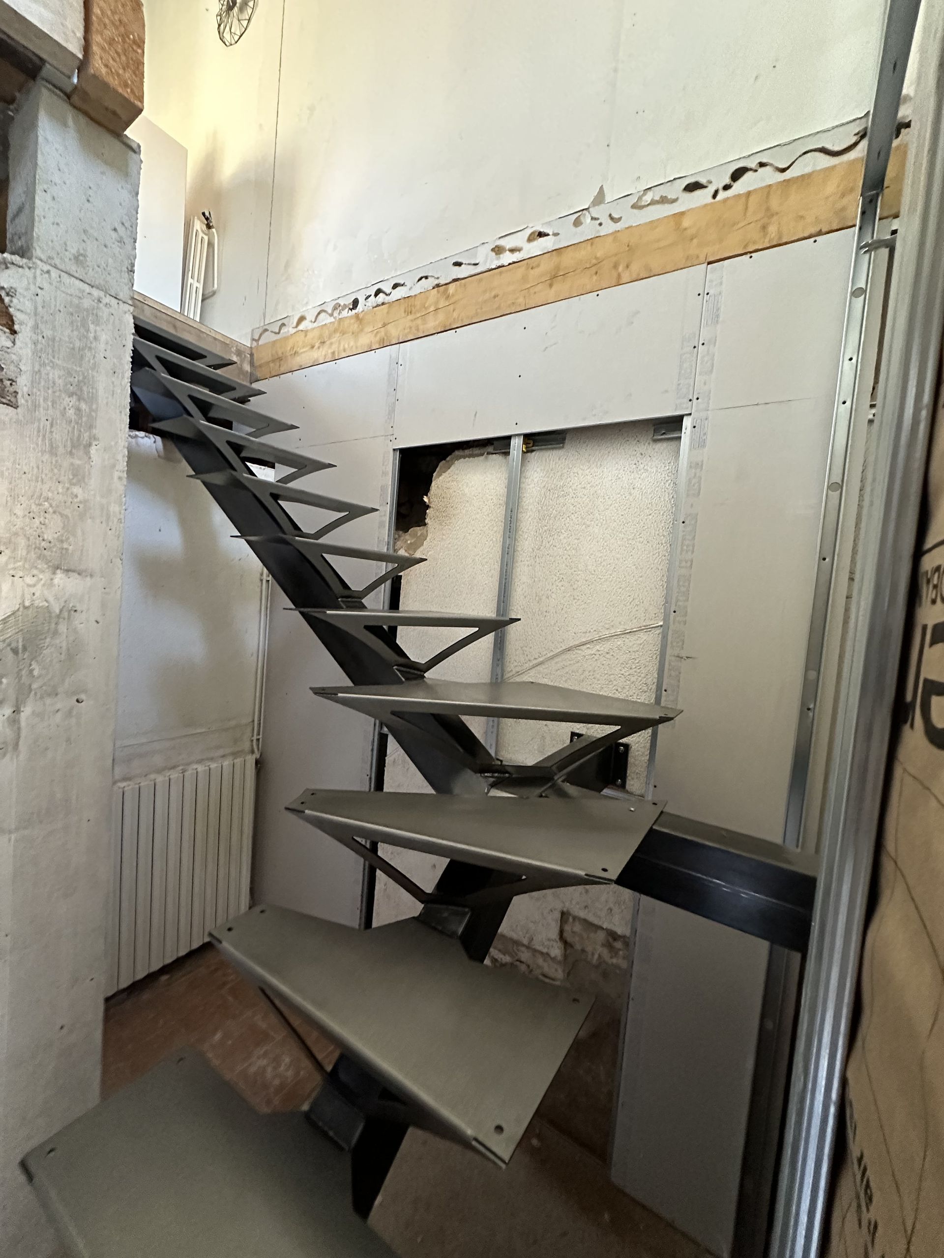 Un escalier métallique moderne, inachevé, avec des marches flottantes, installé à l'intérieur d'une pièce inachevée aux cloisons sèches.