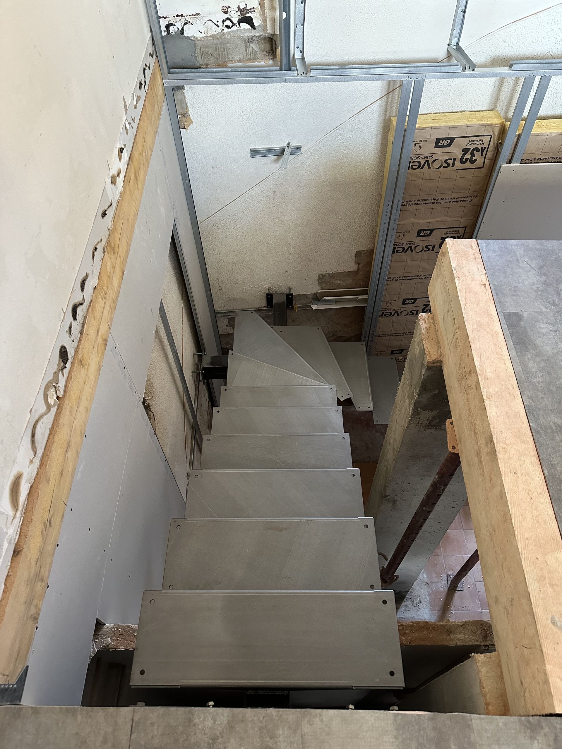 Vue plongeante d'un escalier en bois inachevé menant à un sous-sol partiellement rénové, avec isolation et ossature murale apparentes.