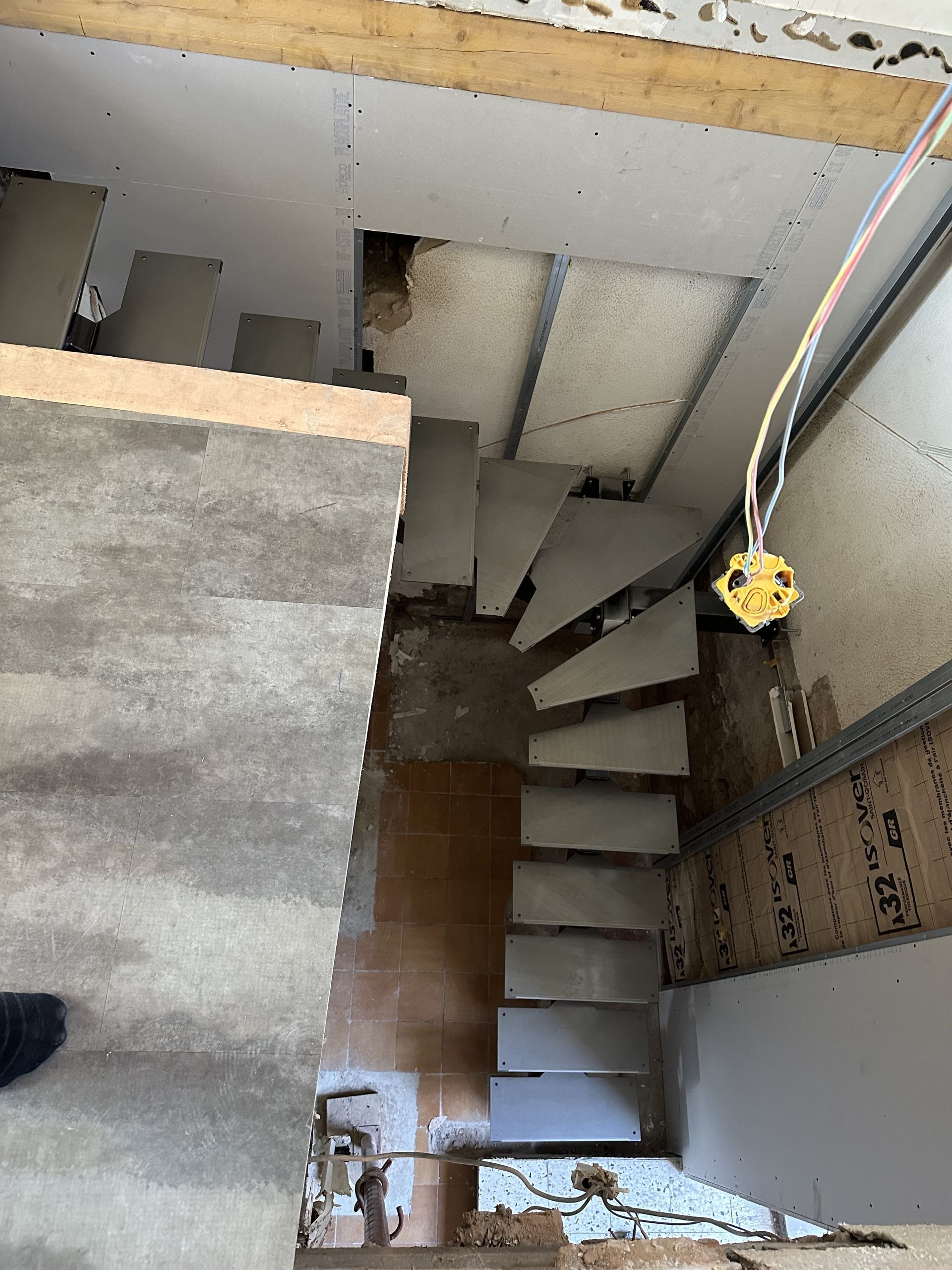 Vue en plongée d'un escalier en bois étroit et inachevé, en construction dans une pièce aux murs nus et aux cloisons sèches.
