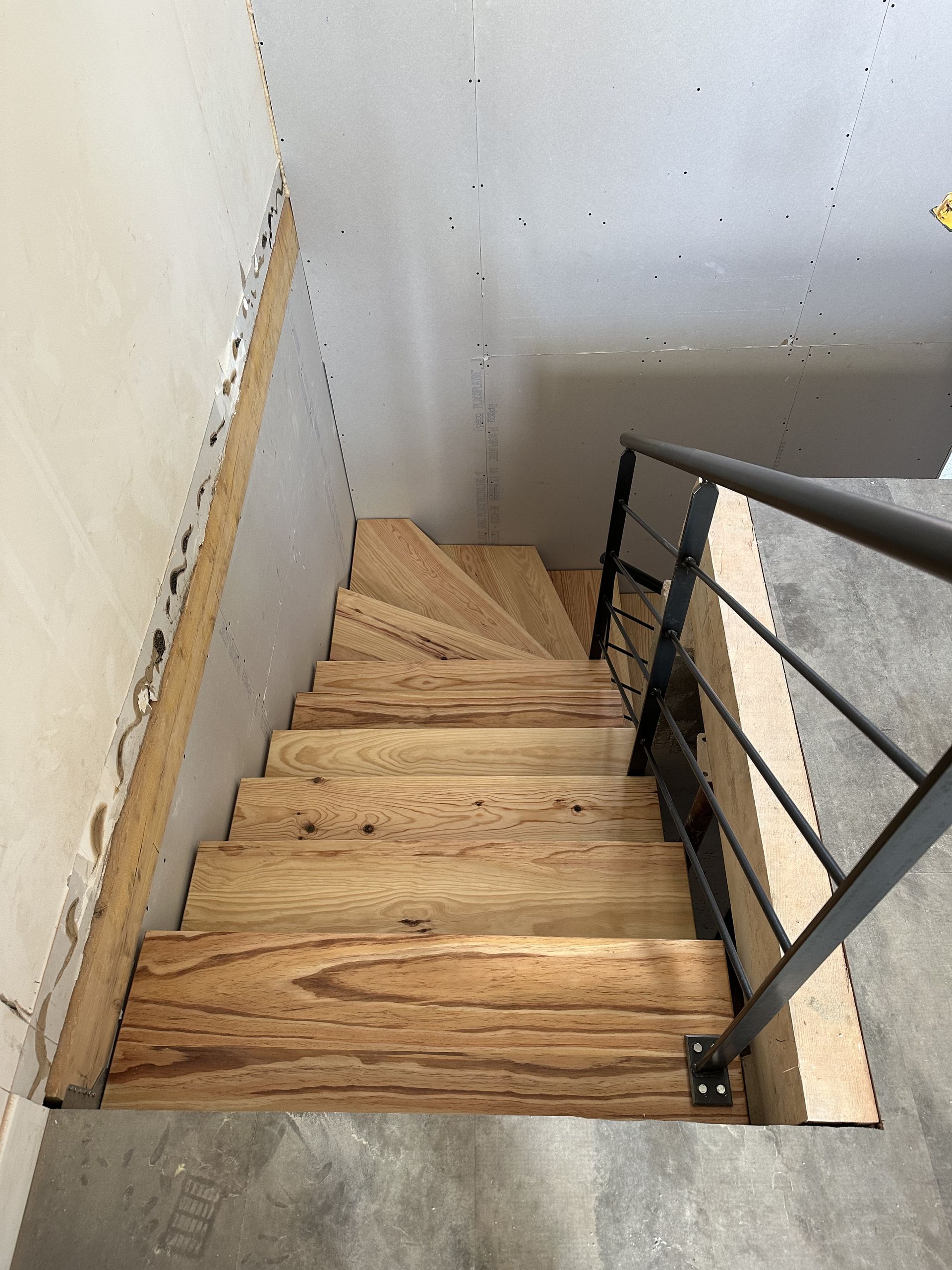 Vue plongeante d'un escalier en bois brut avec une rampe métallique, descendant dans une pièce aux murs en plaques de plâtre grises.