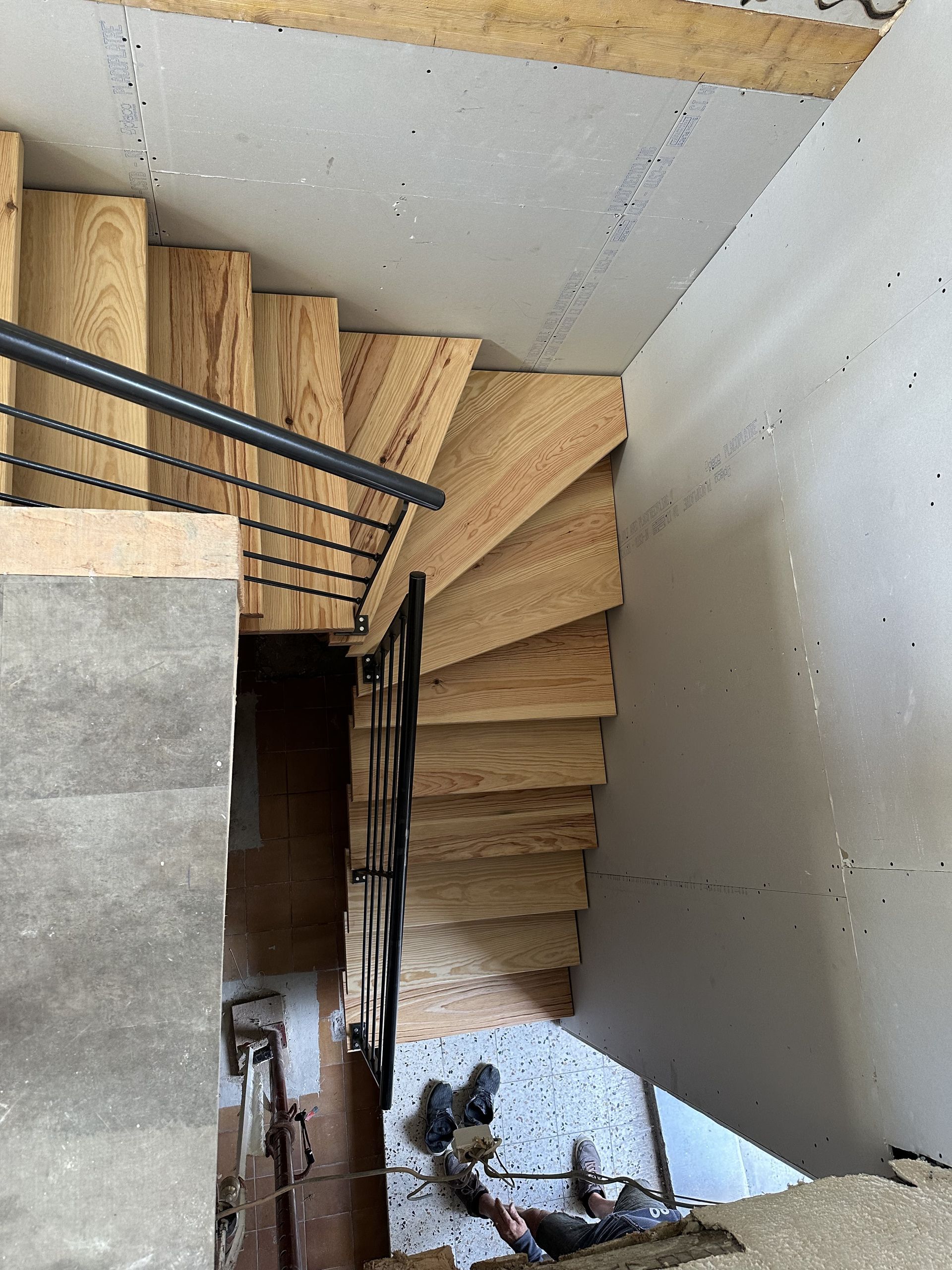 Vue plongeante d'un escalier en bois inachevé avec une rampe en métal noir en cours de construction, donnant sur un sol carrelé.