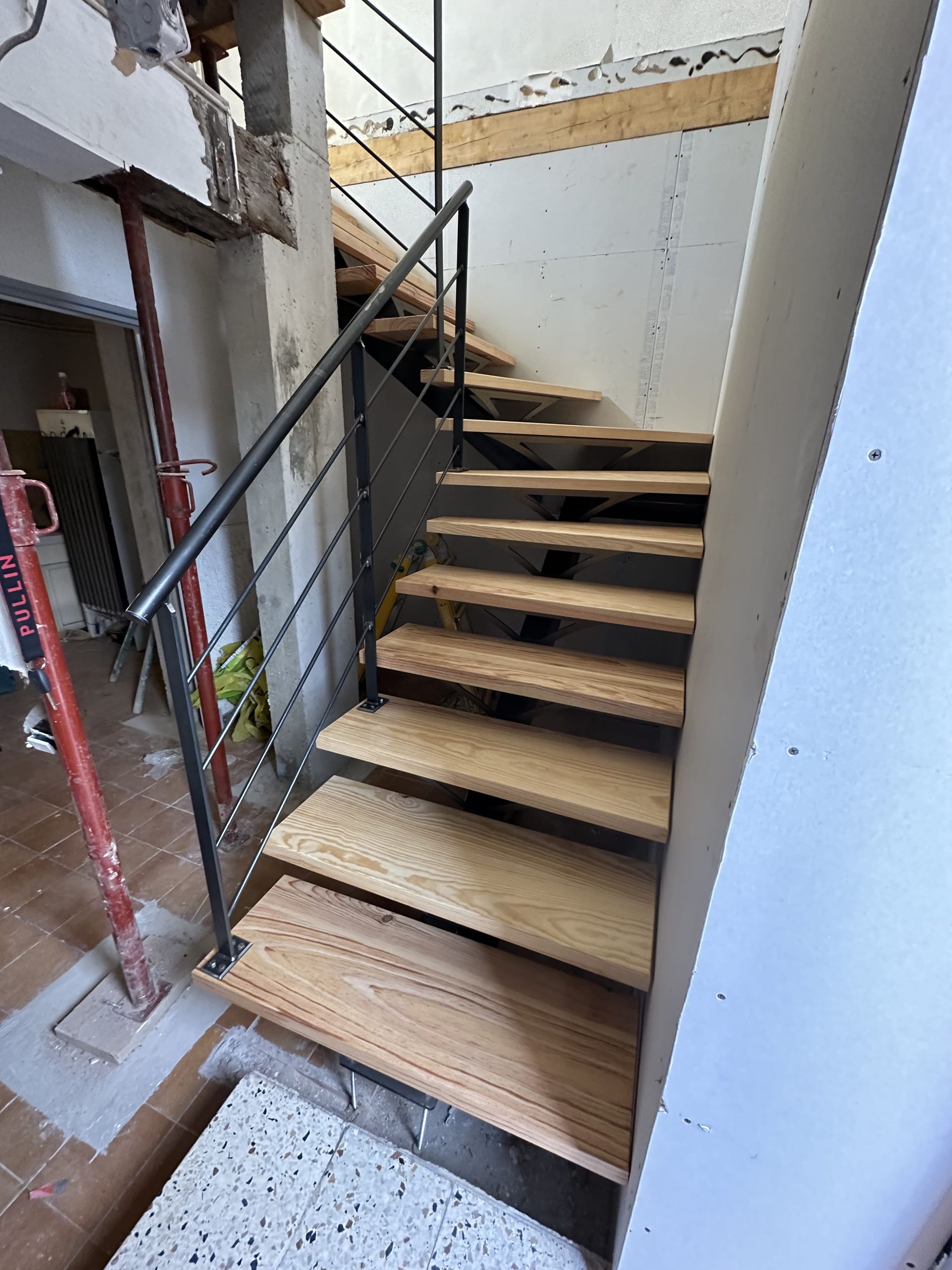 Escalier en bois avec rampes en métal noir dans une pièce en construction, laissant apparaître les murs et les poutres structurelles.