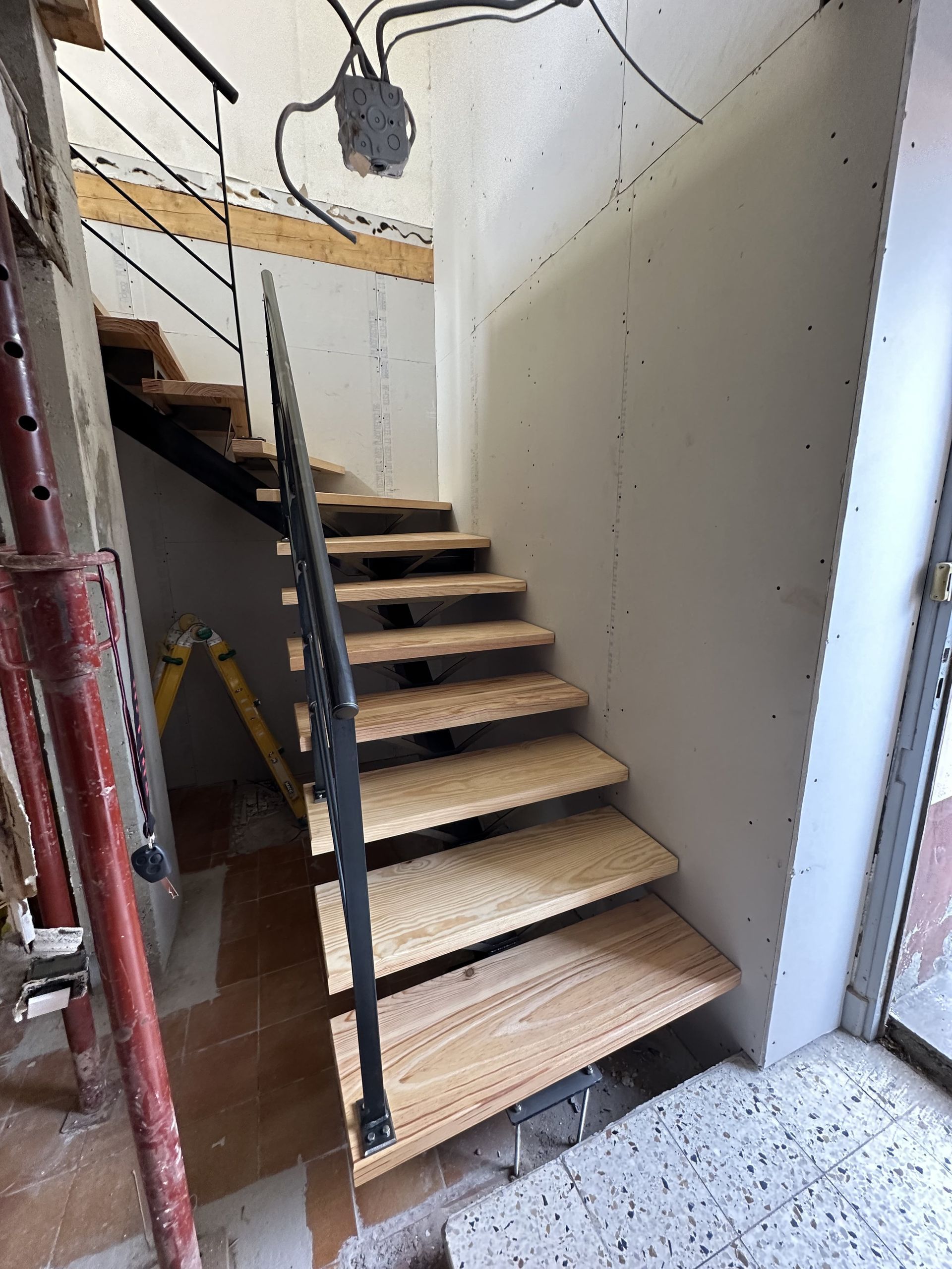 Un escalier en bois inachevé avec une rampe en métal noir, à l'intérieur d'une pièce en construction aux murs blancs.