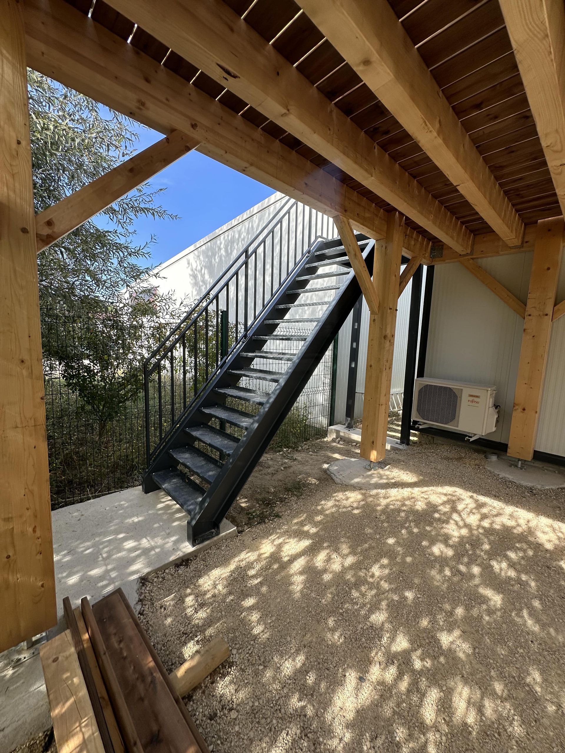 Escalier extérieur en métal sous une terrasse en bois, adossé à un mur blanc, avec un sol en gravier et un climatiseur.