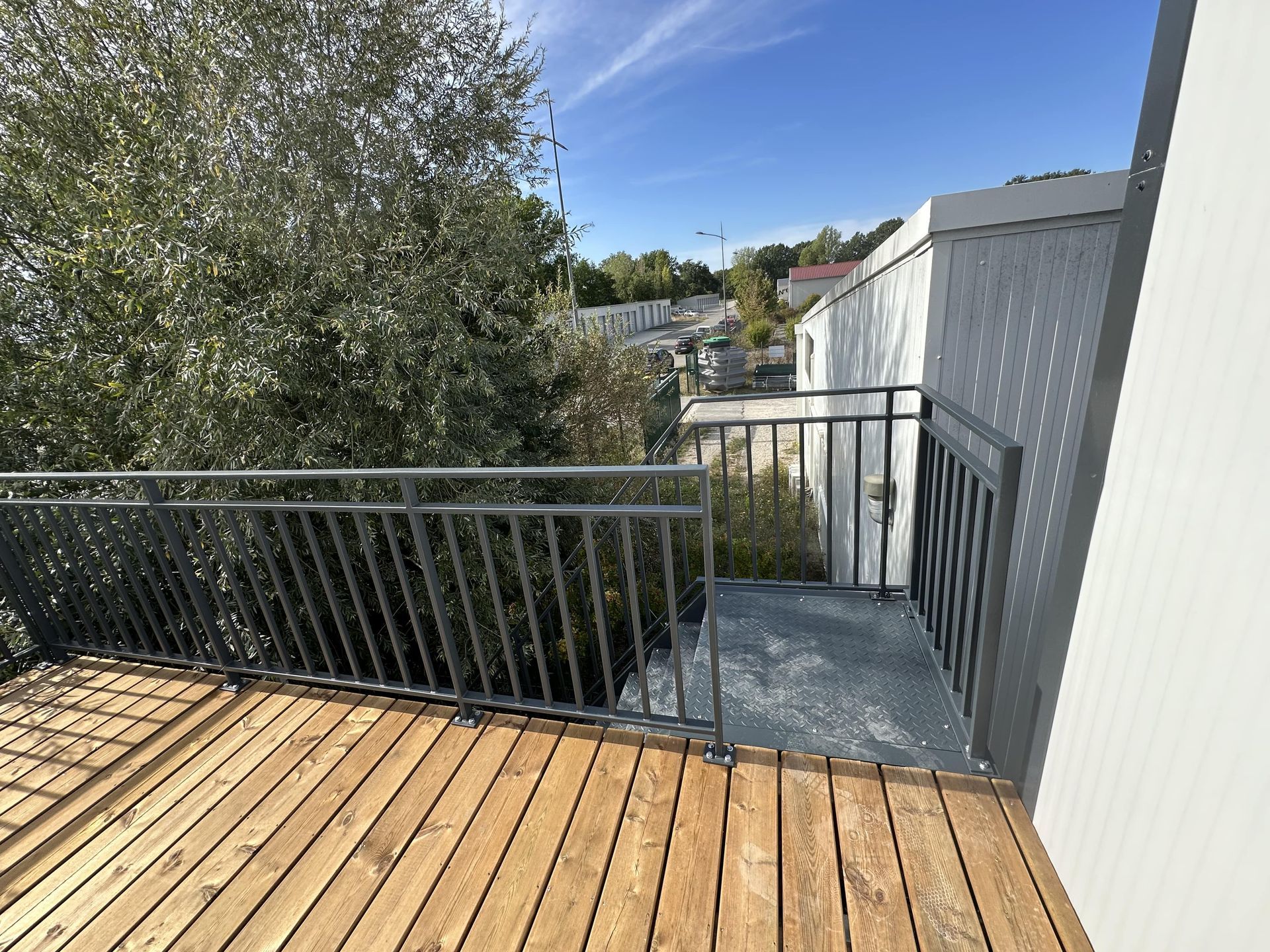 Une terrasse en bois avec des rambardes métalliques surplombe des arbres et un bâtiment blanc sous un ciel bleu ensoleillé.