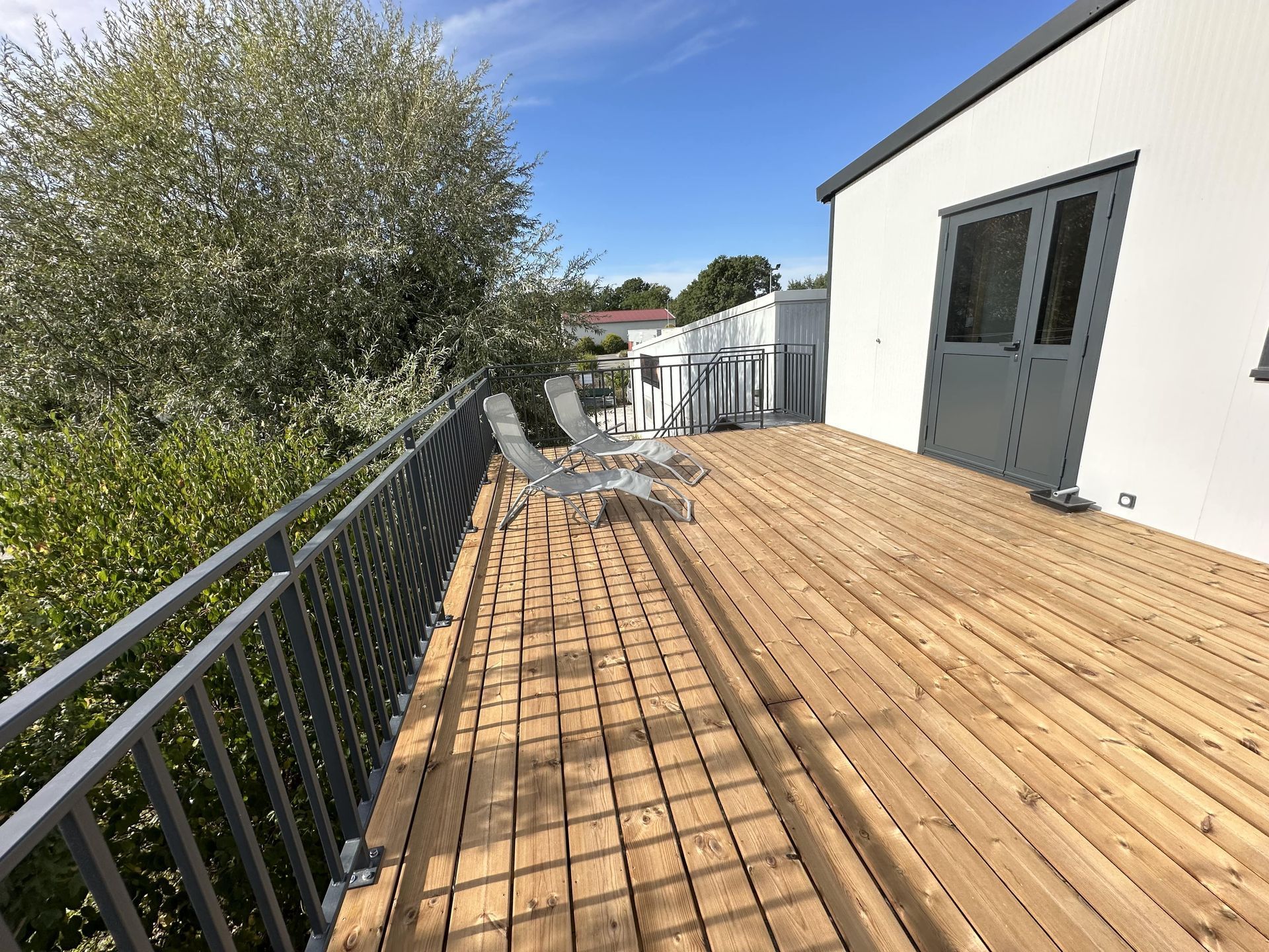Une terrasse en bois avec deux chaises longues, jouxtant un bâtiment blanc et un arbre feuillu, le tout entouré d'une rambarde en métal gris.