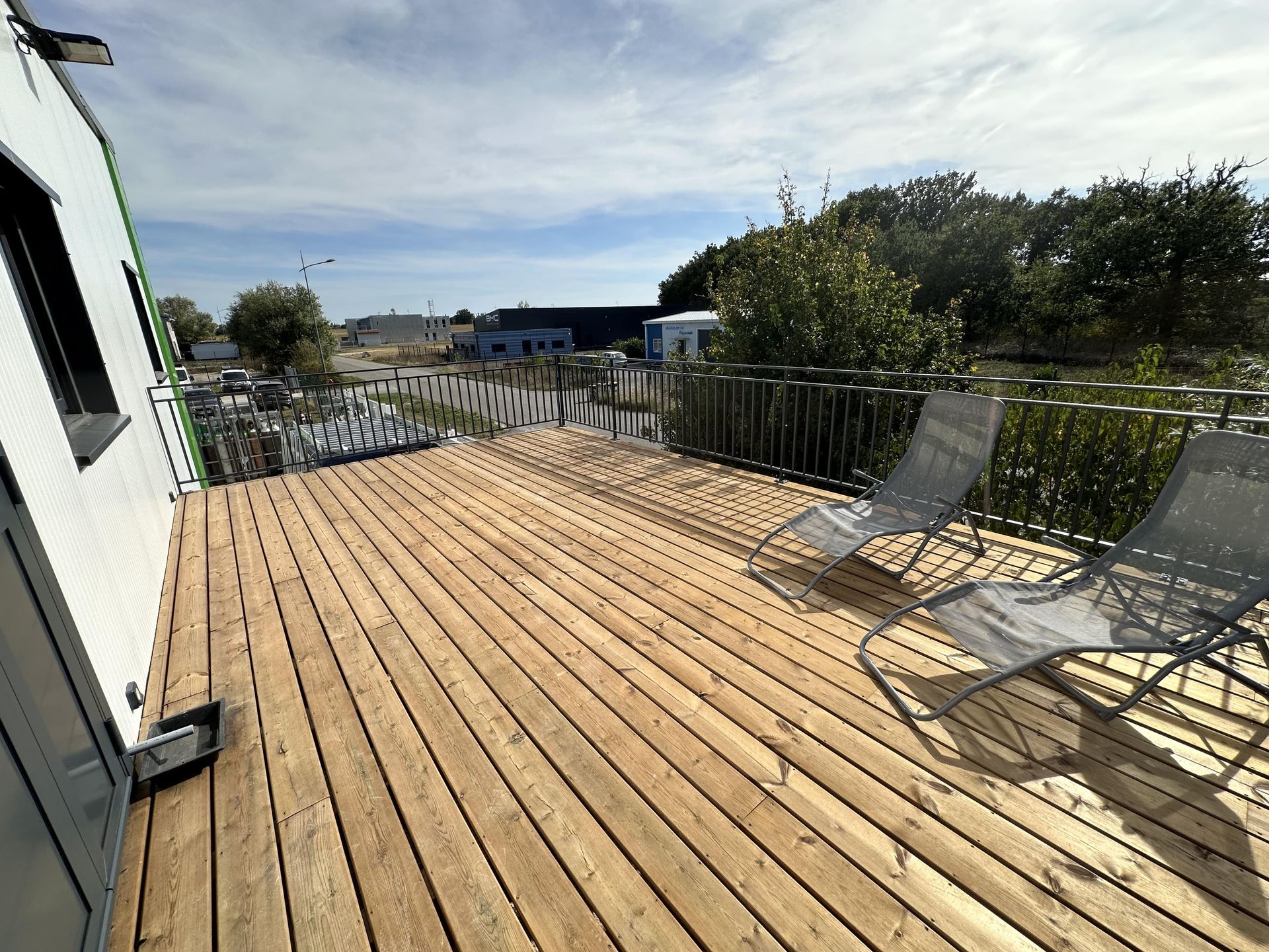 Deux chaises longues grises sont installées sur une terrasse en bois clair donnant sur un espace clôturé avec des arbres et des bâtiments sous un ciel nuageux.