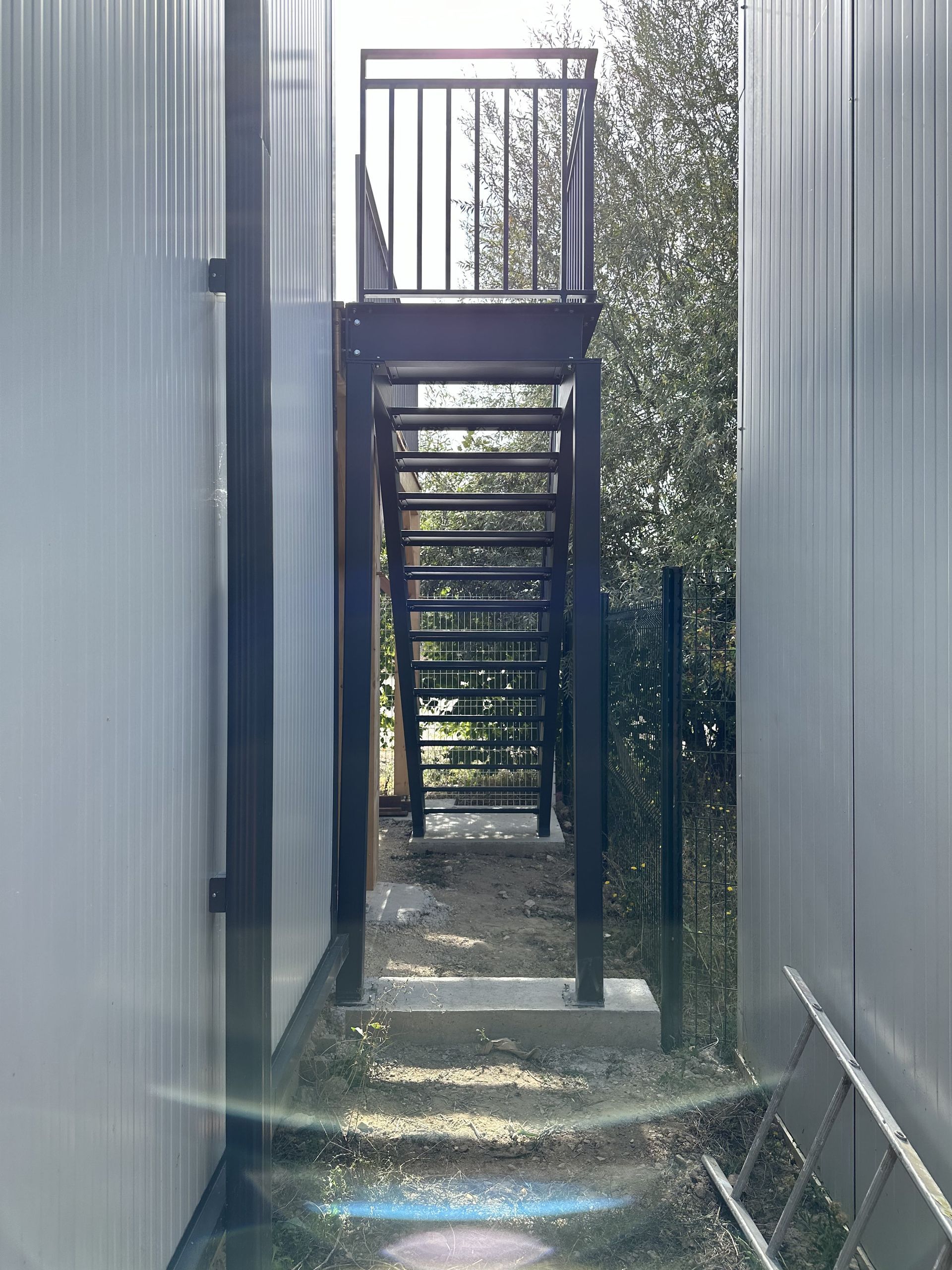 Un escalier extérieur en métal, entre deux parois métalliques, mène à une plateforme surélevée où filtre la lumière naturelle à travers les arbres.