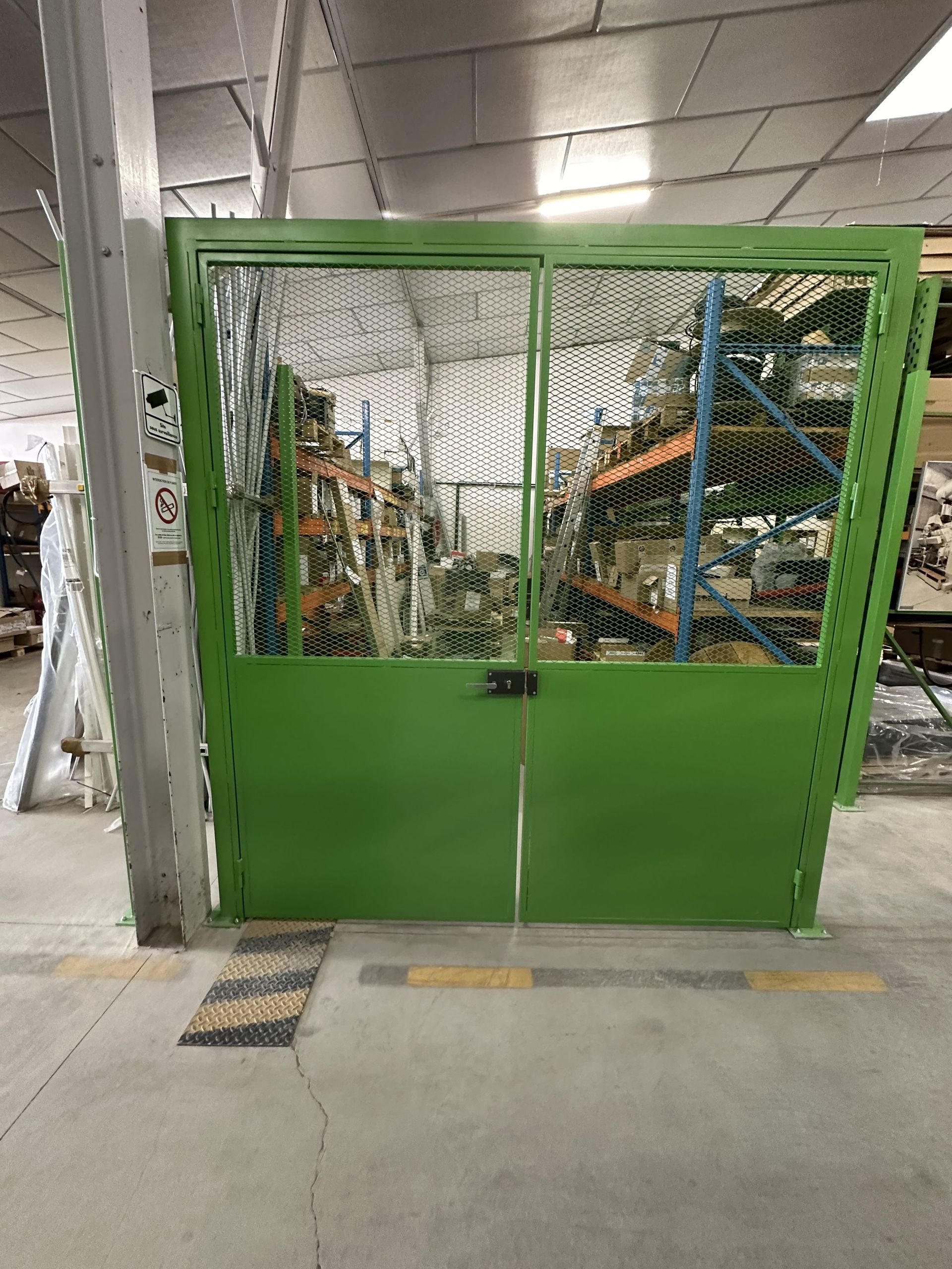 Une barrière de sécurité en métal vert, surmontée d'un treillis métallique, se trouve dans une zone de stockage d'un entrepôt industriel.