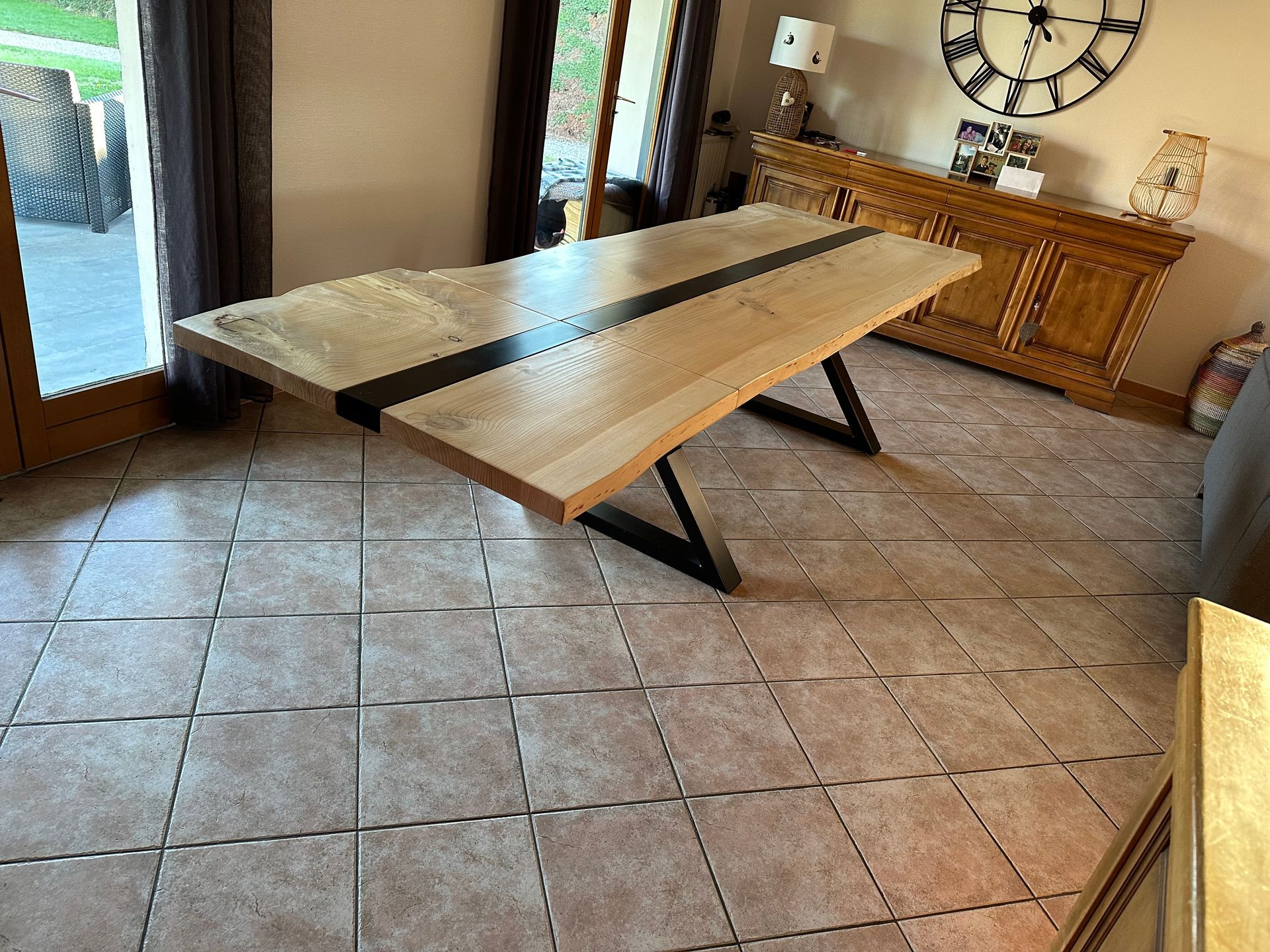 Une grande table à manger en bois avec une incrustation centrale noire et des pieds angulaires en métal noir, dans une pièce carrelée et ensoleillée.