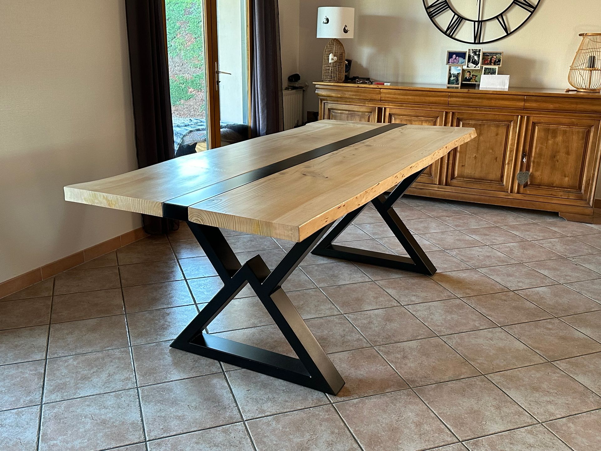 Une table à manger en bois clair avec une incrustation centrale foncée et des pieds en métal noir en forme de X dans une pièce meublée.