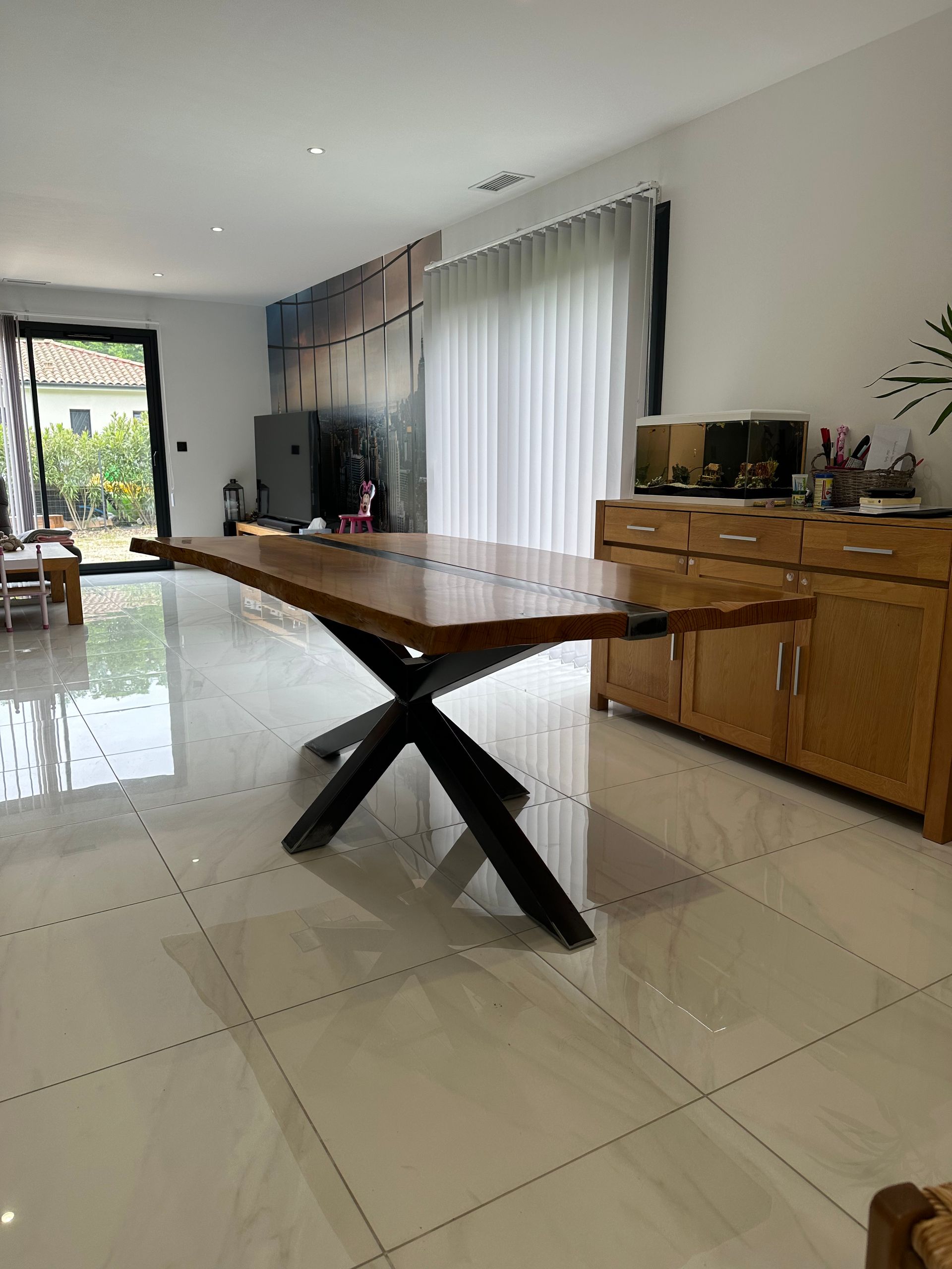 Une table à manger en bois brut avec un piètement en X en métal noir, dans une salle à manger lumineuse carrelée avec un buffet en bois.
