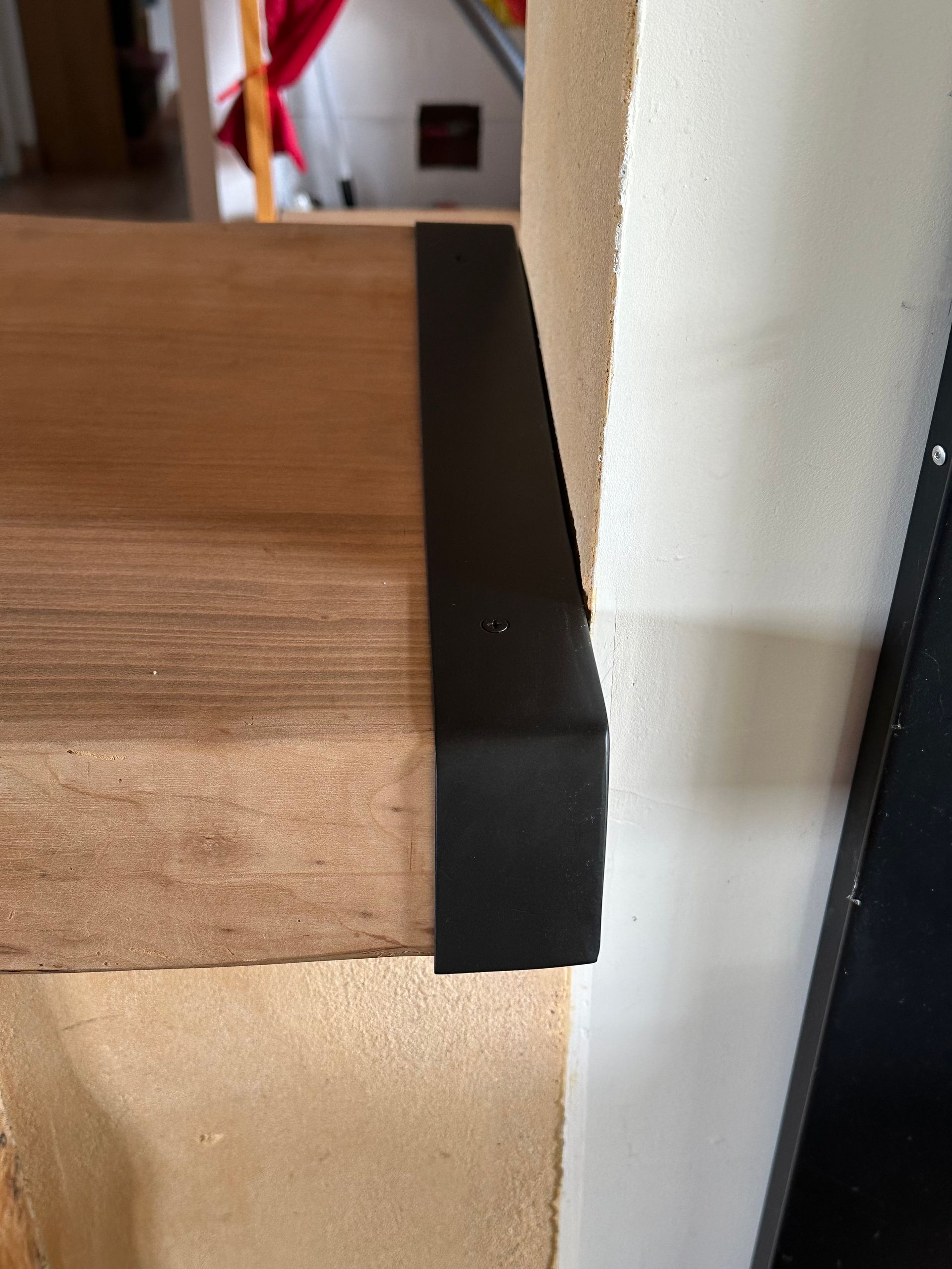 Un protège-bord en métal noir est installé sur le coin d'une étagère en bois clair, contre un mur blanc texturé.