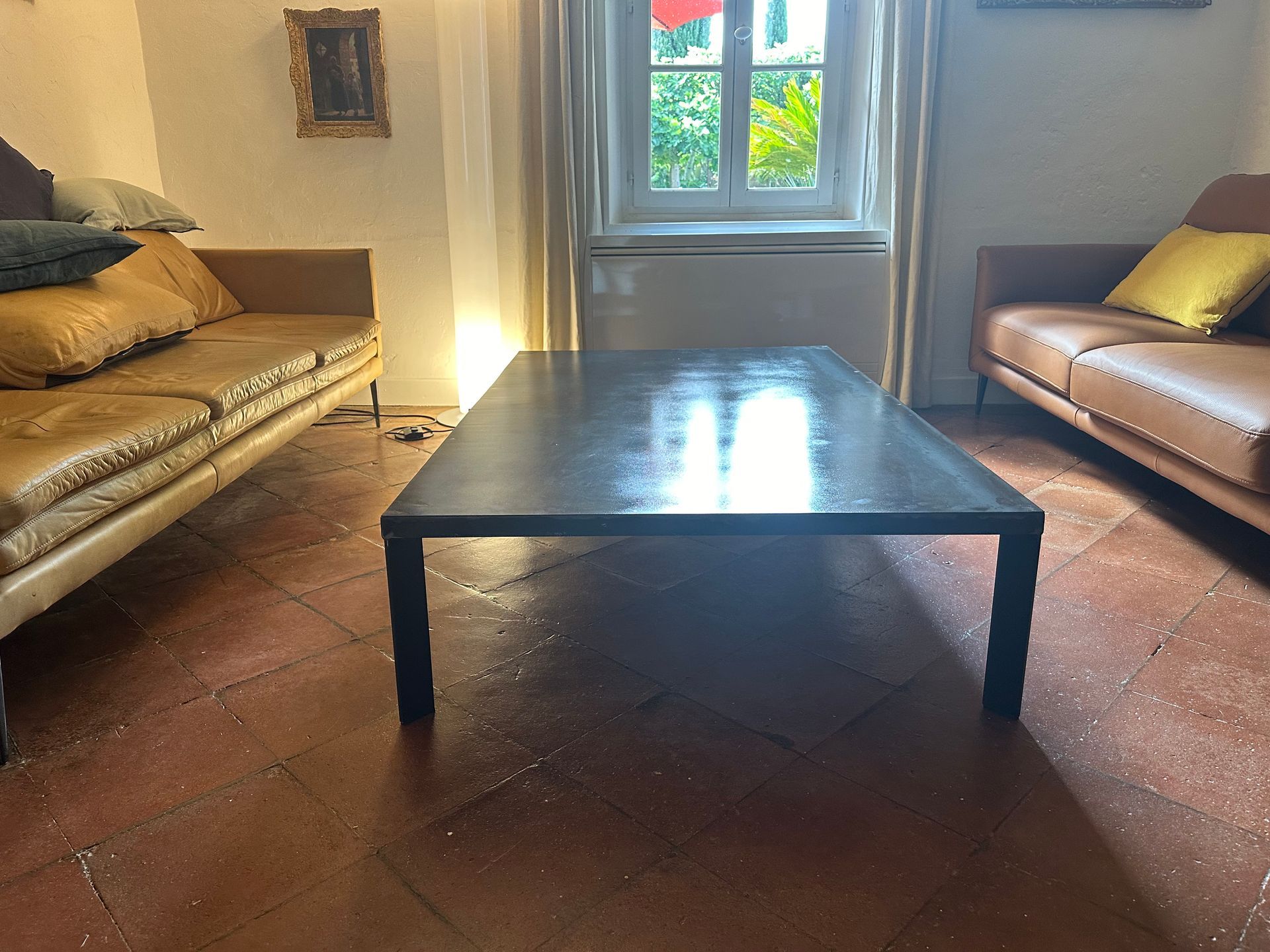 Dans un salon au sol carrelé de terre cuite, une table basse sombre est placée entre deux canapés en cuir beige.