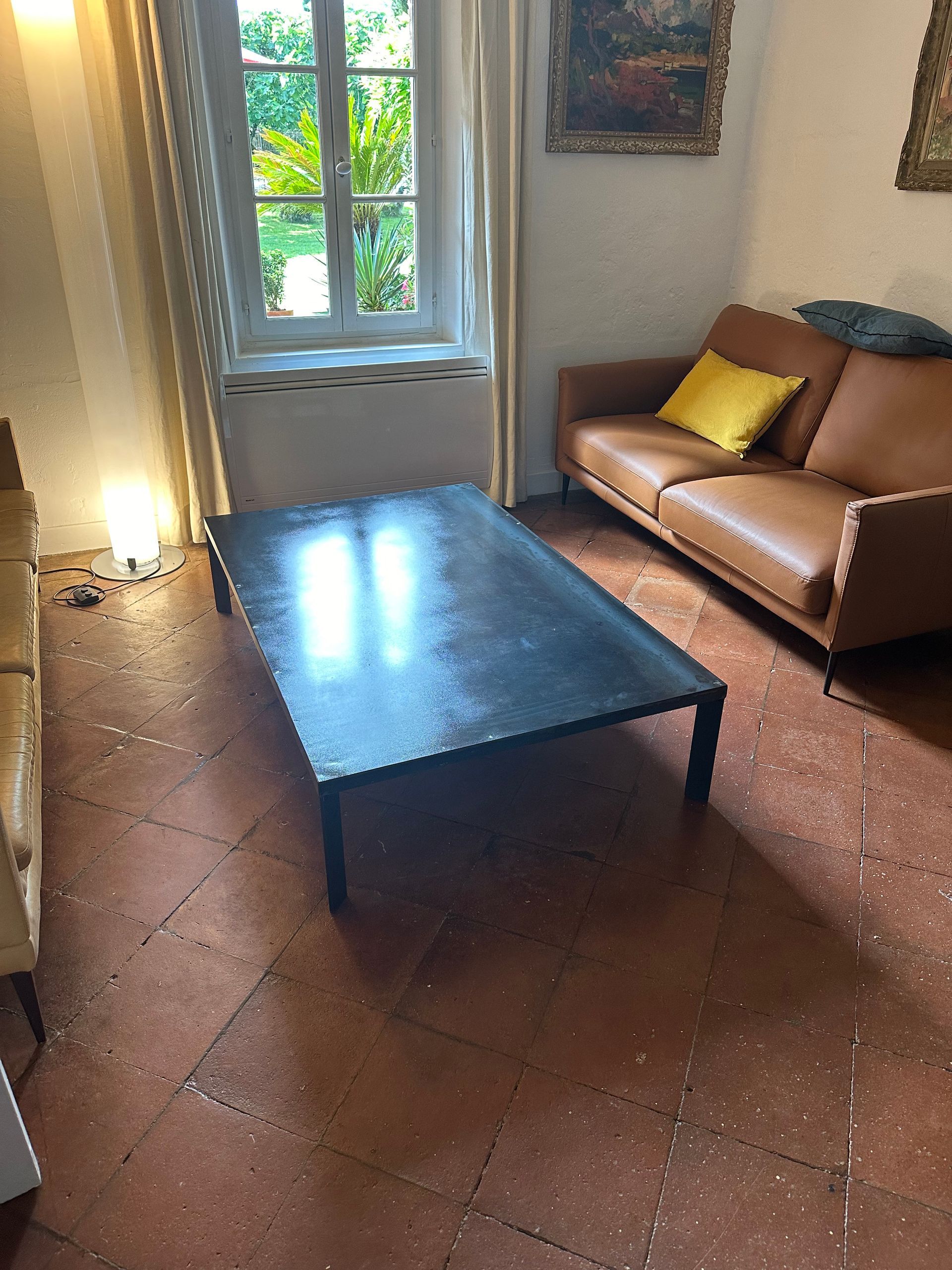 Un canapé en cuir marron avec un coussin jaune est placé près d'une fenêtre dans une pièce avec une table basse noire et un sol en terre cuite.