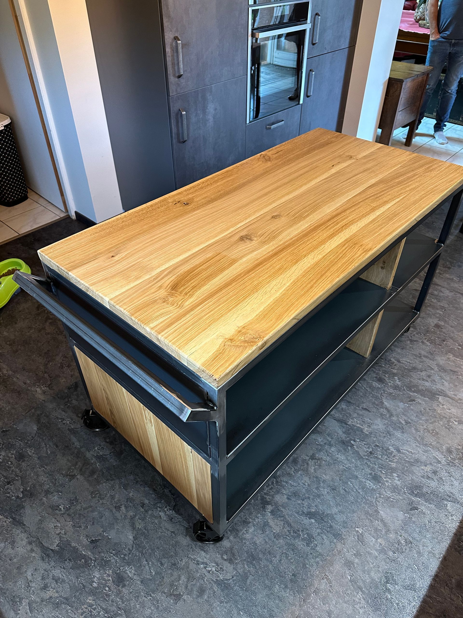 Un îlot de cuisine rectangulaire avec un plateau en bois clair façon bloc de boucher et une structure en métal gris foncé avec des étagères sur roulettes.