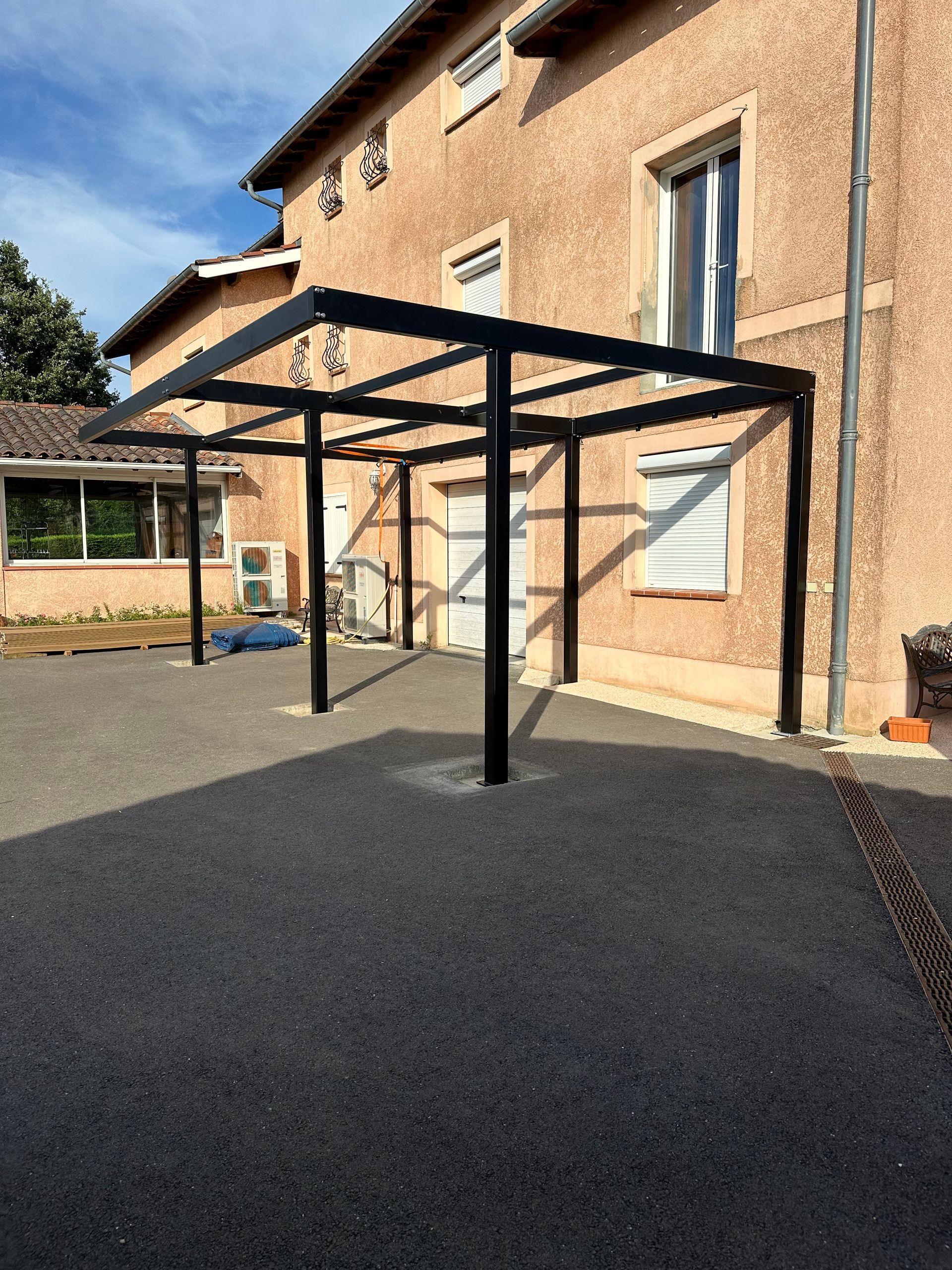 Une structure d'abri de voiture en acier noir se dresse sur une allée de gravier devant une maison beige à plusieurs étages.