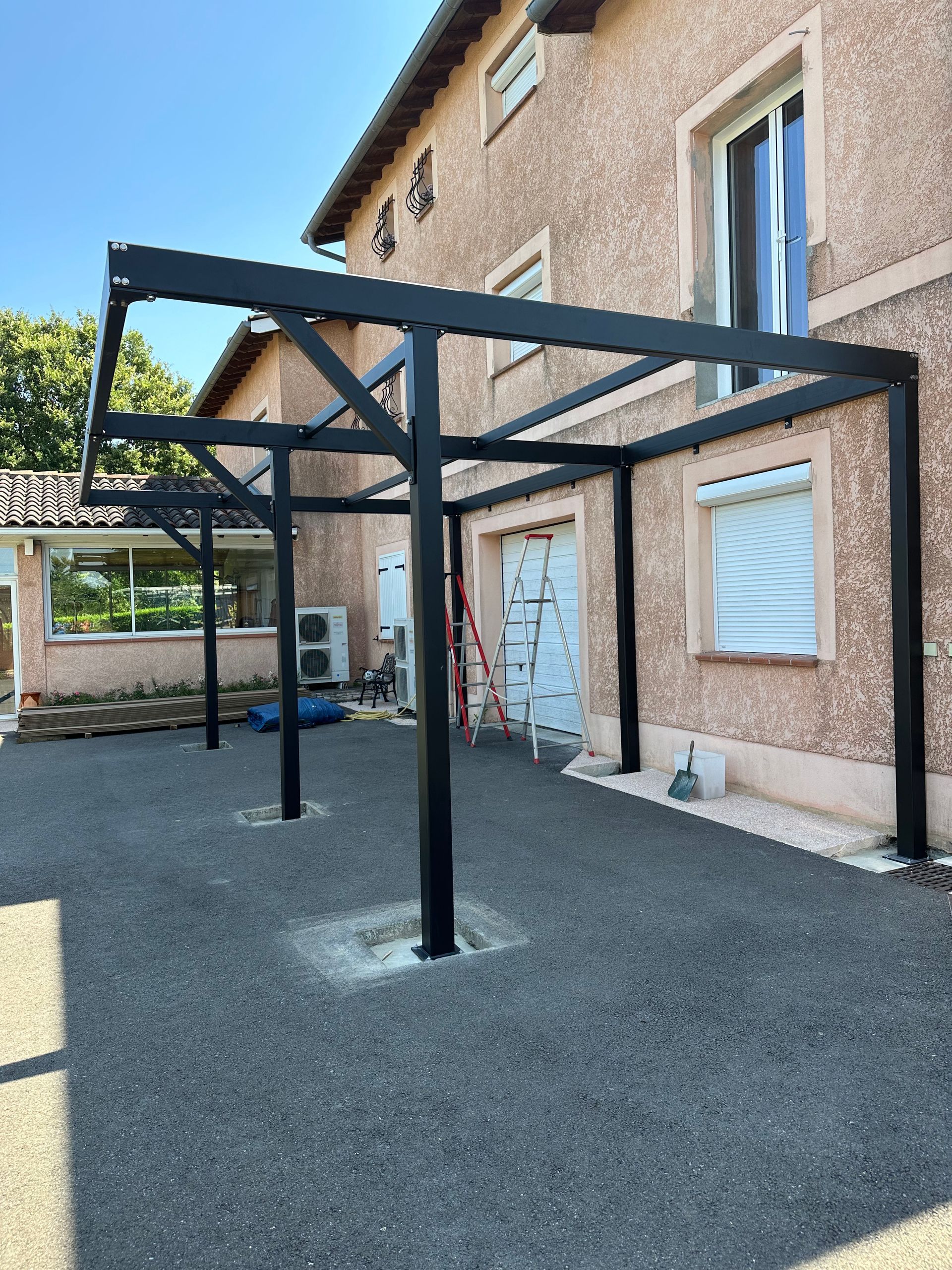 Une structure métallique noire pour une couverture de terrasse est installée contre une maison en stuc beige, au-dessus d'une allée de gravier gris.