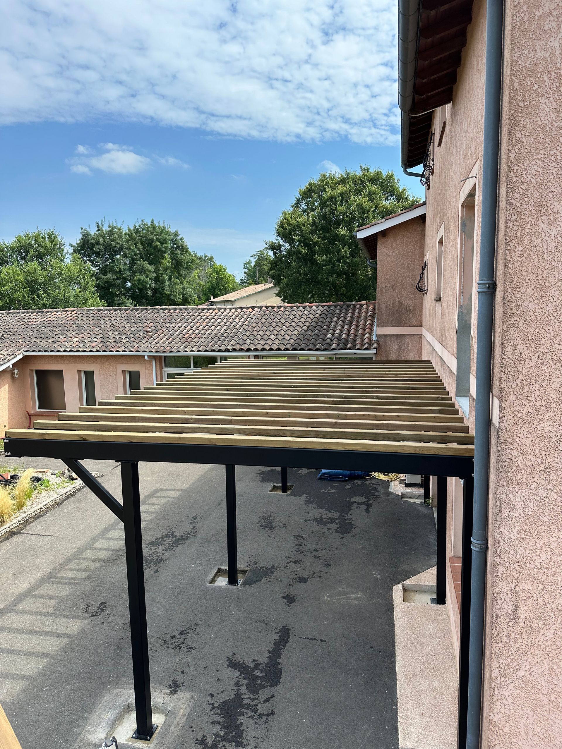 Une pergola en bois nouvellement construite, avec des supports en métal noir, s'étendant sur le côté d'un bâtiment en stuc brun clair.