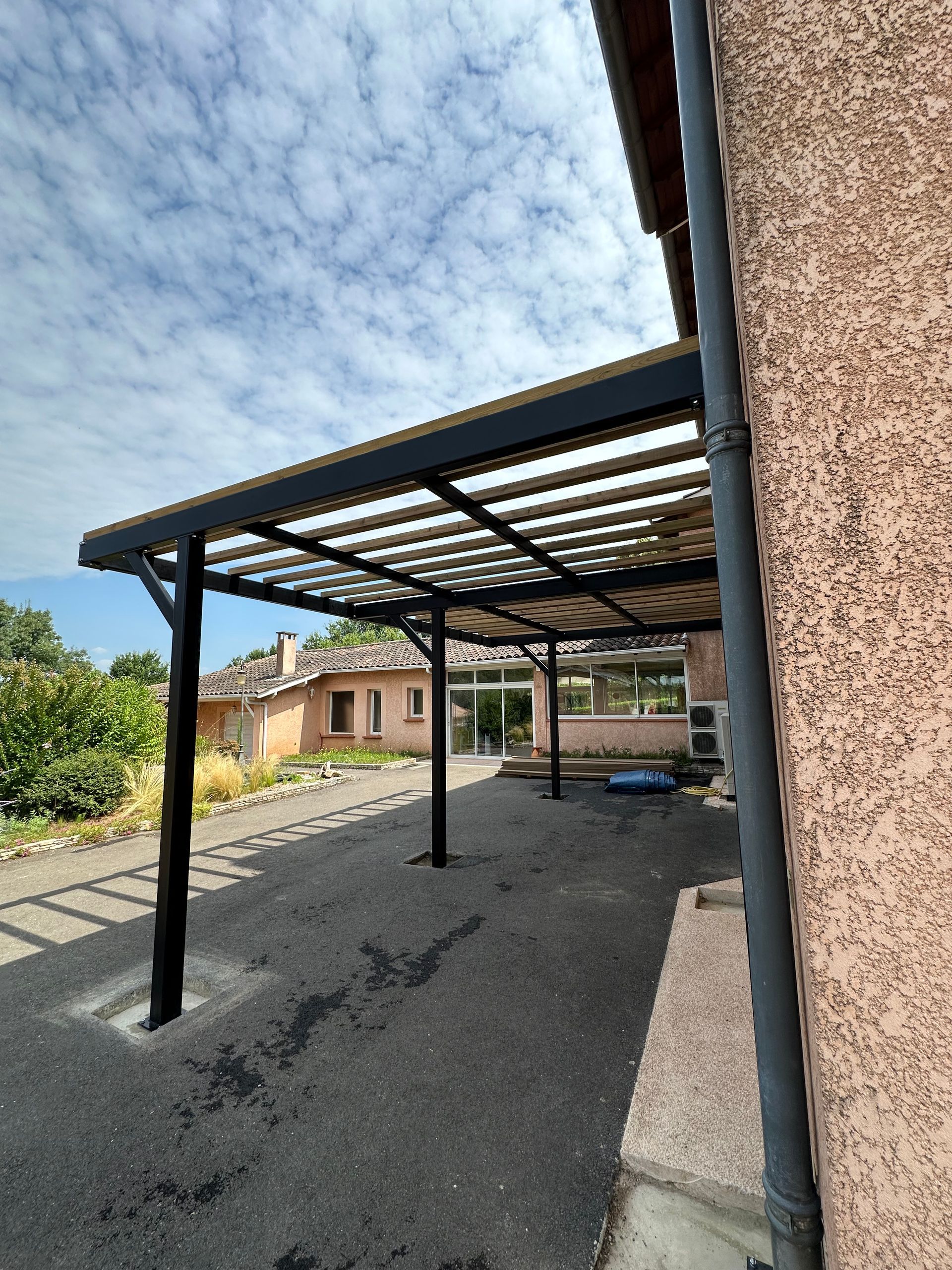 Un abri de voiture en métal noir surplombe une allée de gravier, à côté d'une maison aux murs en stuc, sous un ciel bleu nuageux.