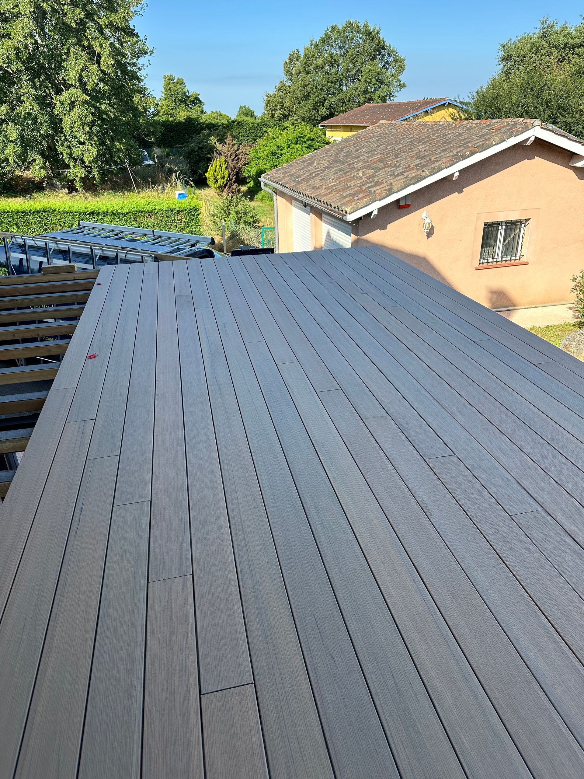Vue en plongée d'une terrasse composite grise en construction, montrant des planches parallèles menant à une maison.