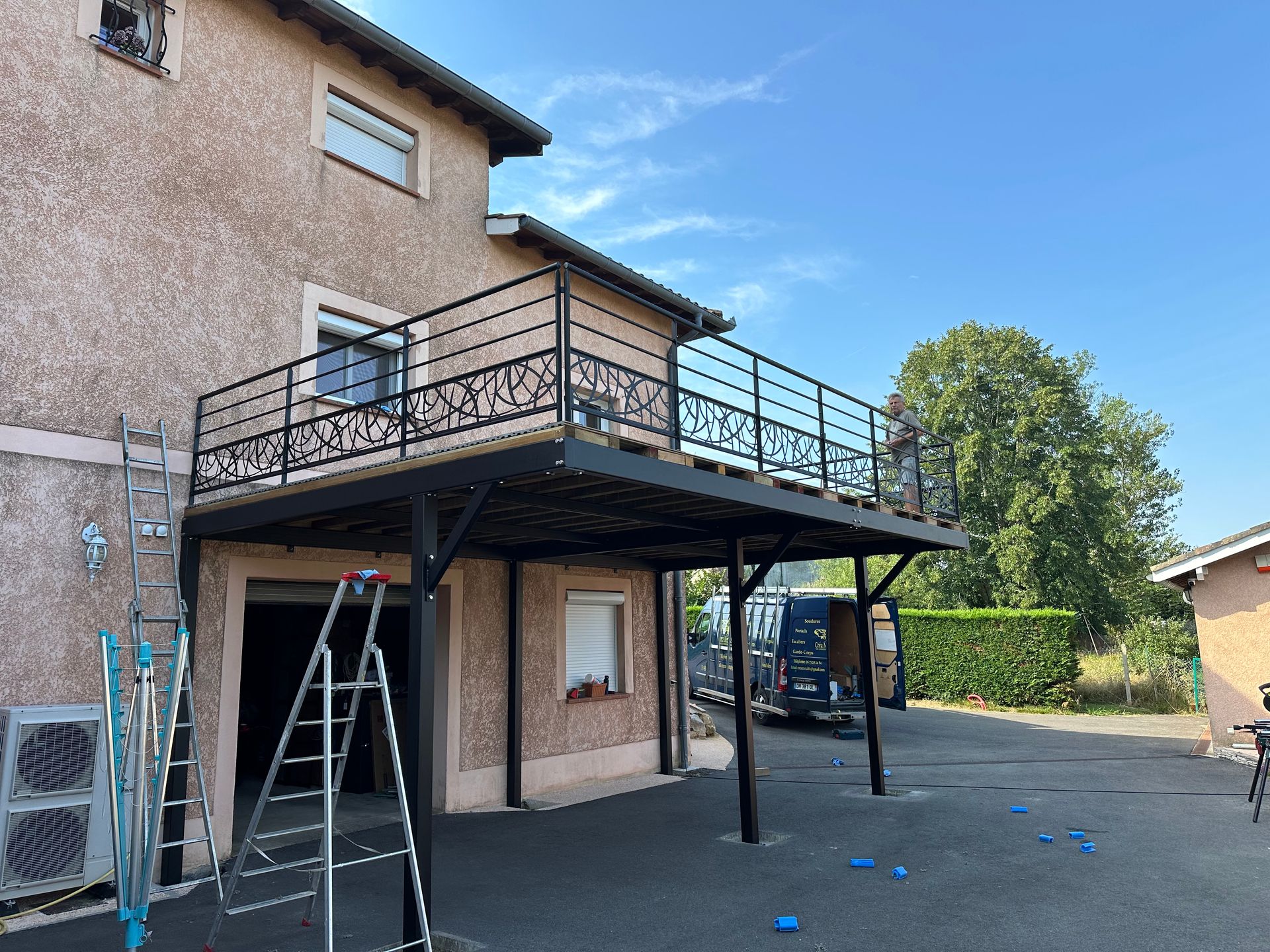 Une maison avec un balcon en métal noir récemment installé et des piliers de soutien, avec des échelles et une camionnette de chantier dans l'allée.
