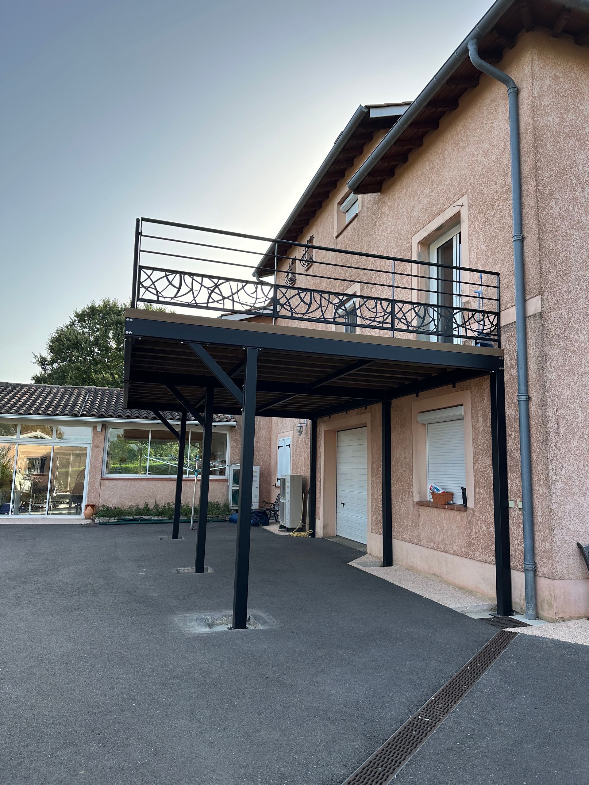 Une maison de deux étages en stuc rose avec un balcon en métal noir et un abri voiture couvert au-dessus d'une allée pavée.