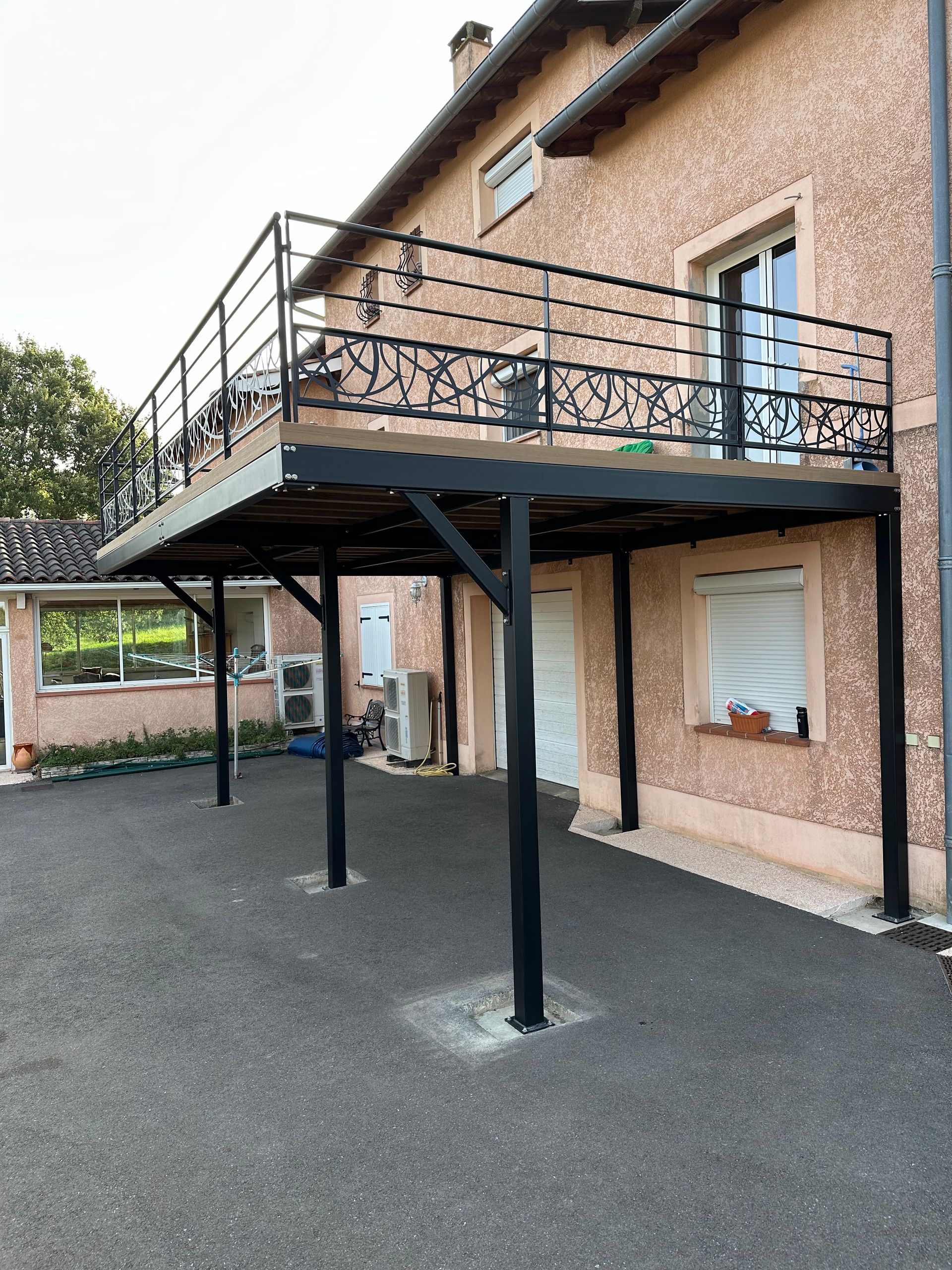 Une grande structure métallique noire abritant un balcon et une voiture, fixée sur le côté d'une maison beige, avec un sol en gravier.