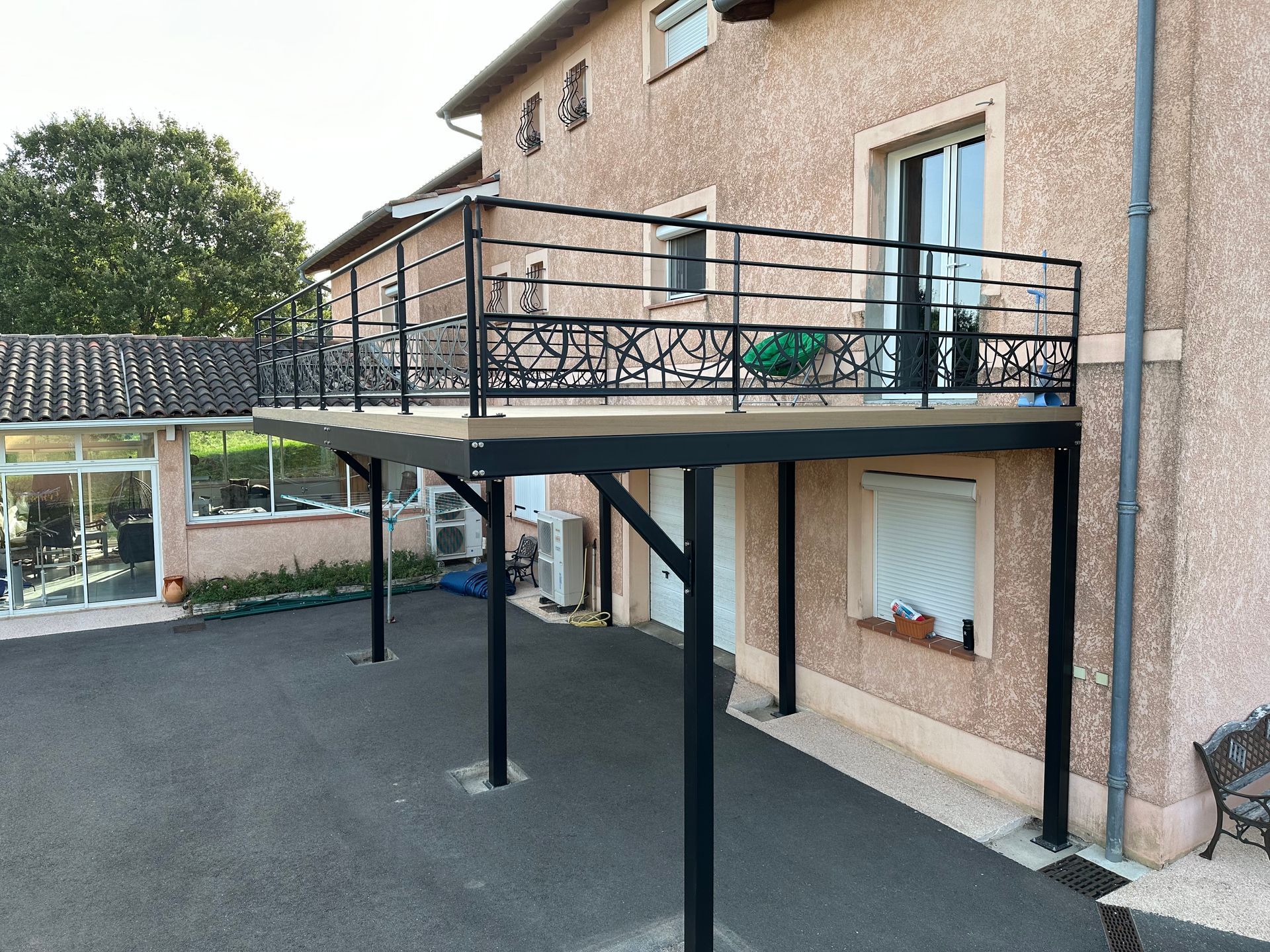 Une maison à l'extérieur beige avec une terrasse surélevée en métal noir et une rambarde soutenue par des piliers au-dessus d'une allée pavée.
