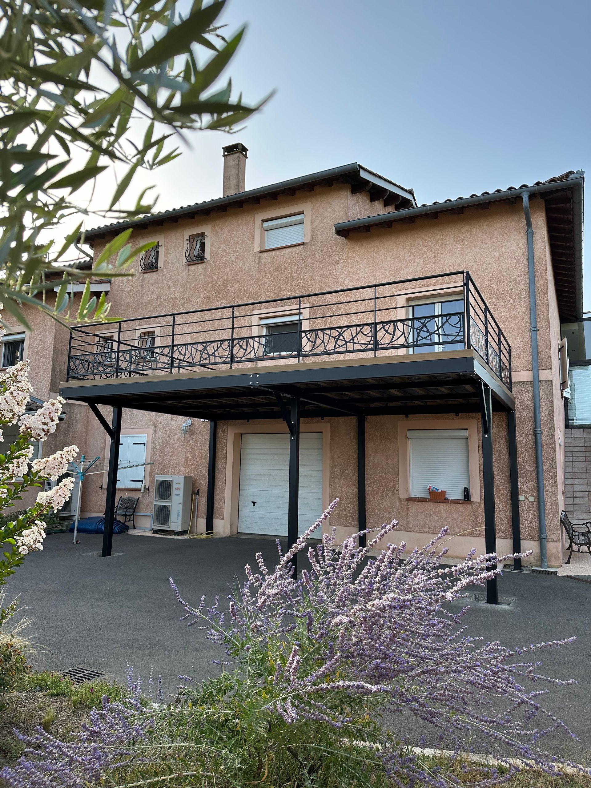 Une maison de trois étages avec un grand balcon en métal noir et un abri voiture, située dans une cour avec des buissons fleuris.