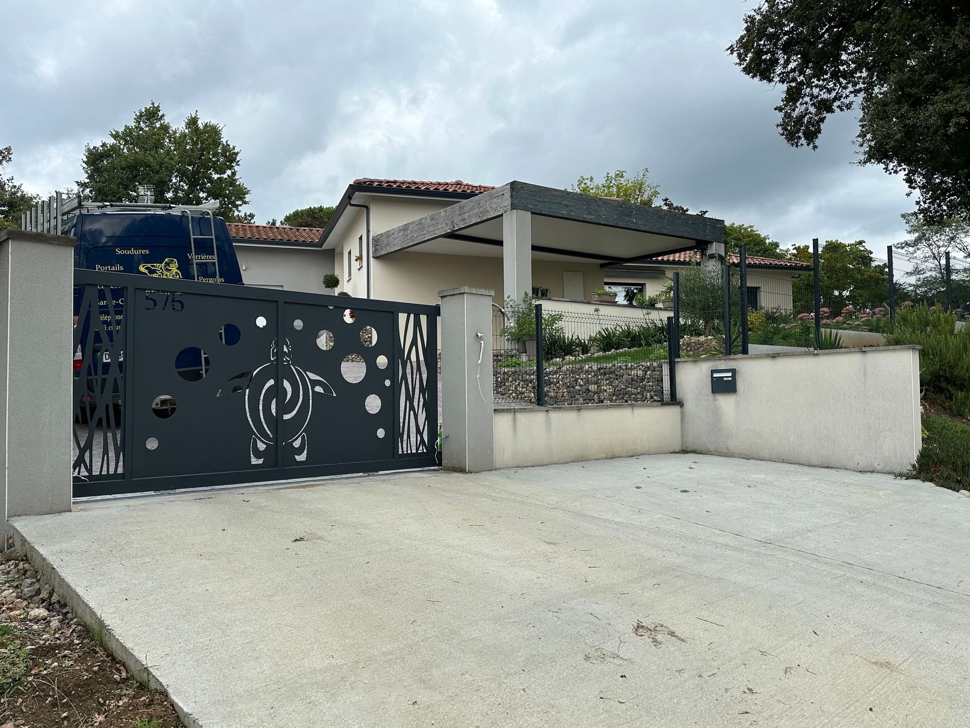 Un portail décoratif gris, orné de découpes circulaires et d'un motif en spirale, se dresse devant une maison moderne dotée d'une allée en béton.