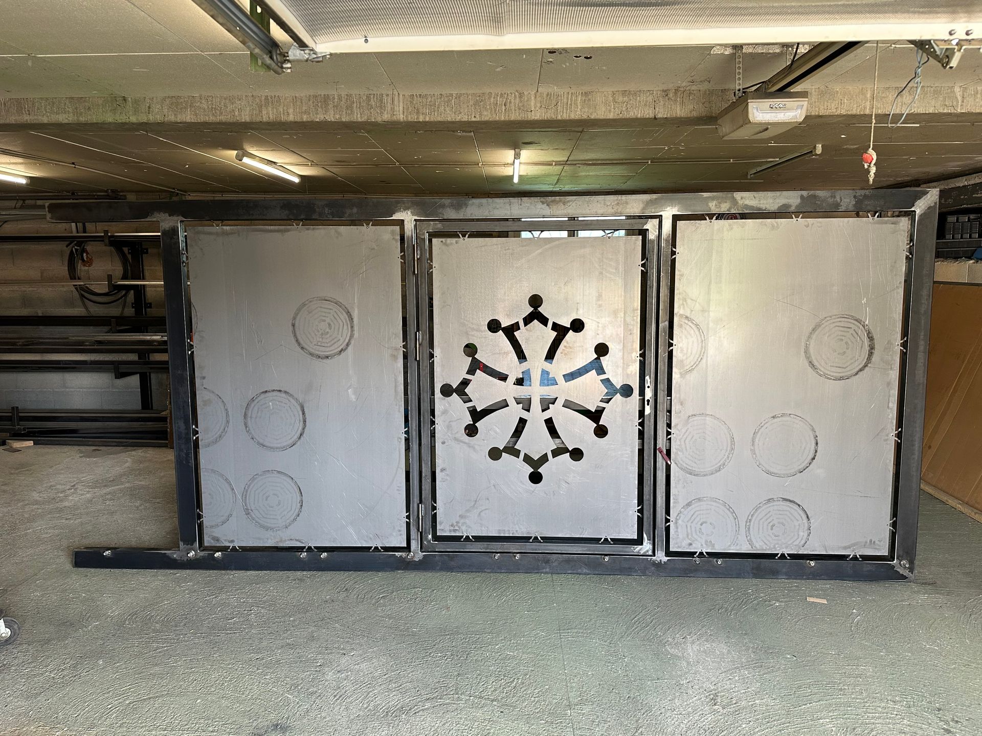 Portail métallique à trois panneaux en cours de fabrication, présentant une découpe centrale en forme de croix stylisée dans un décor d'atelier.